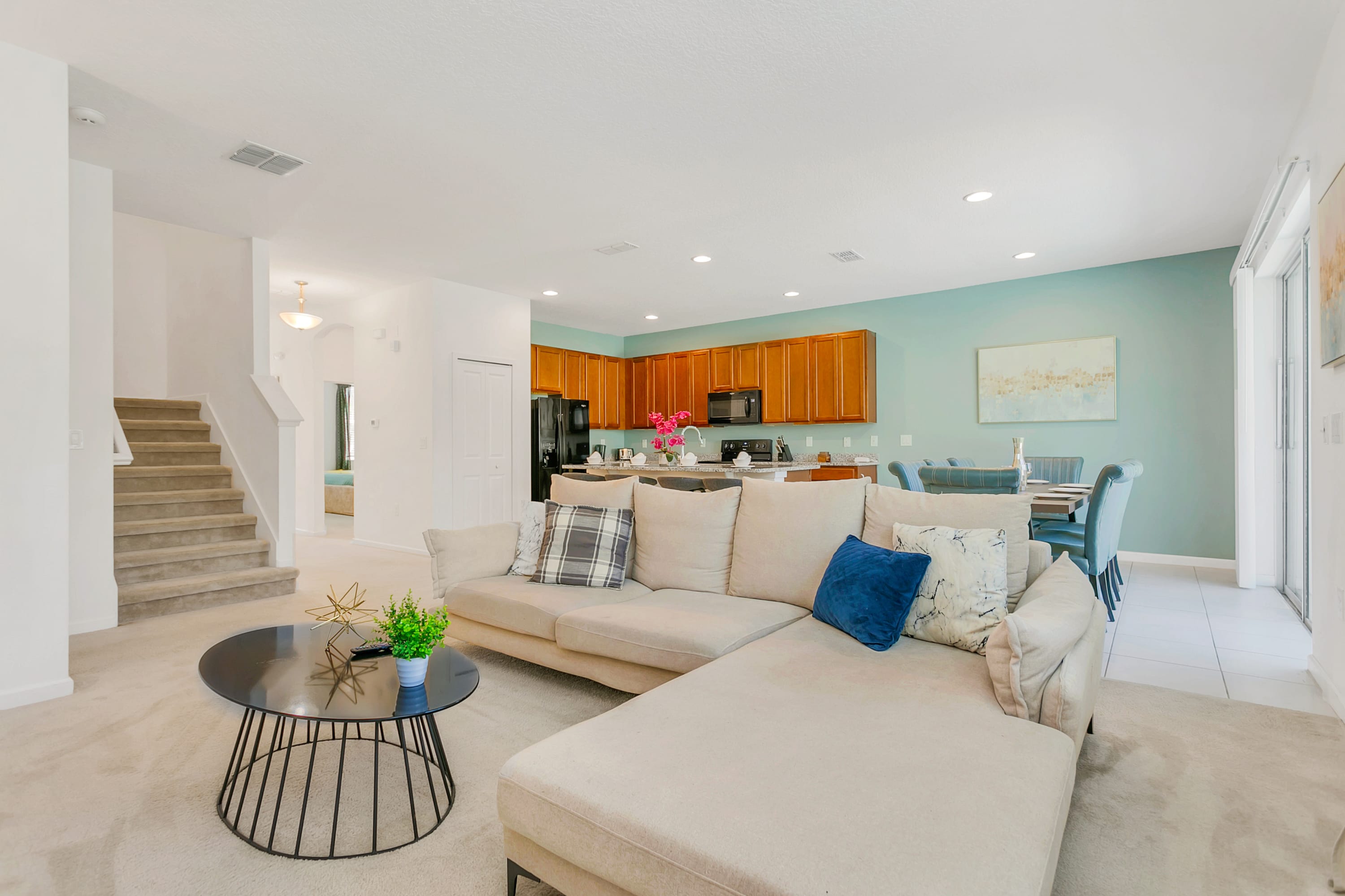 An open-concept living space featuring a large beige sectional sofa with decorative pillows, a black wire-frame coffee table, and a view into the kitchen and dining area. A staircase is visible on the left, and the flooring transitions from carpet to tile.