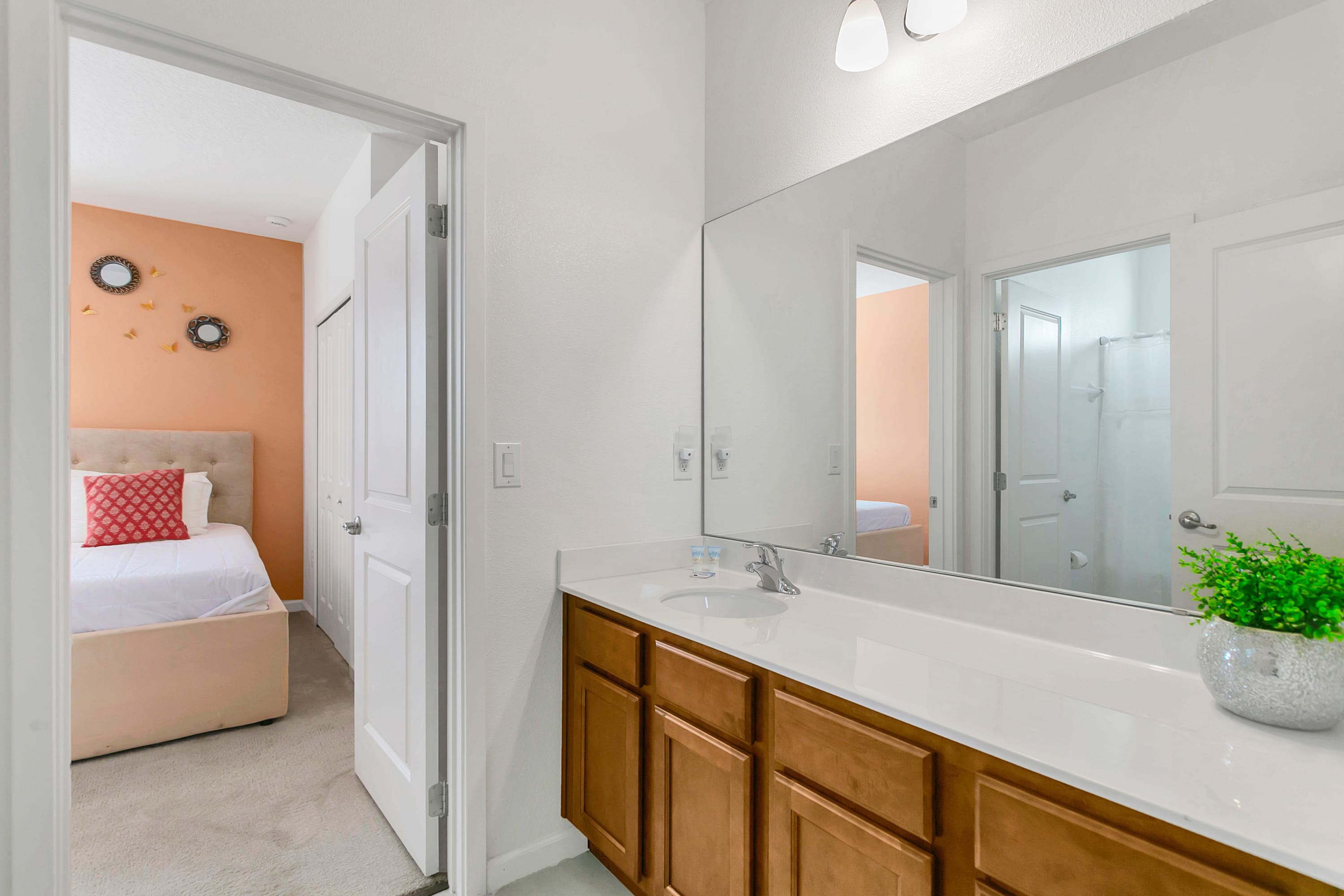 A single vanity bathroom with light wood cabinets and a white countertop, with a large mirror reflecting the doorway to the peach-walled bedroom. A small potted plant sits on the counter.