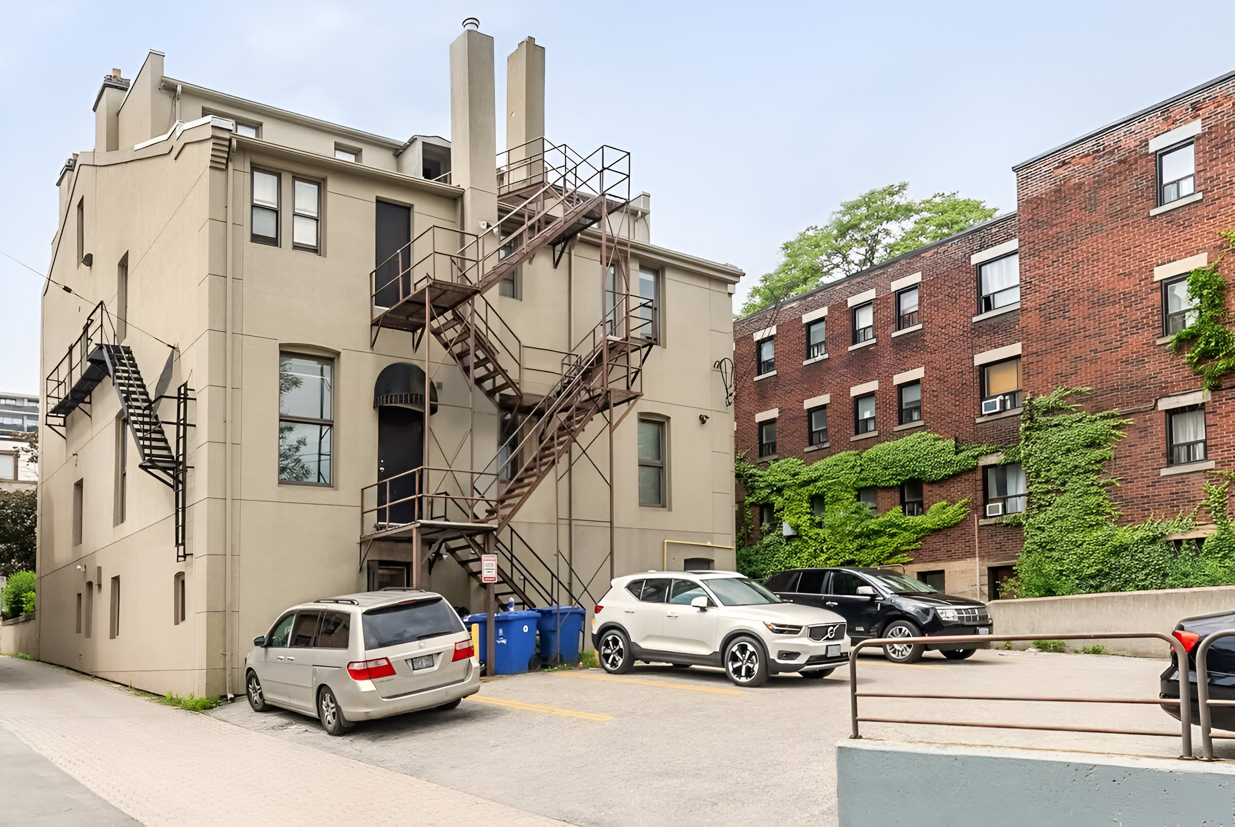 The exterior of a light beige apartment building with multiple fire escapes and windows is shown, alongside a paved parking lot with several cars. A brick building covered in ivy is visible in the background, suggesting an urban setting.