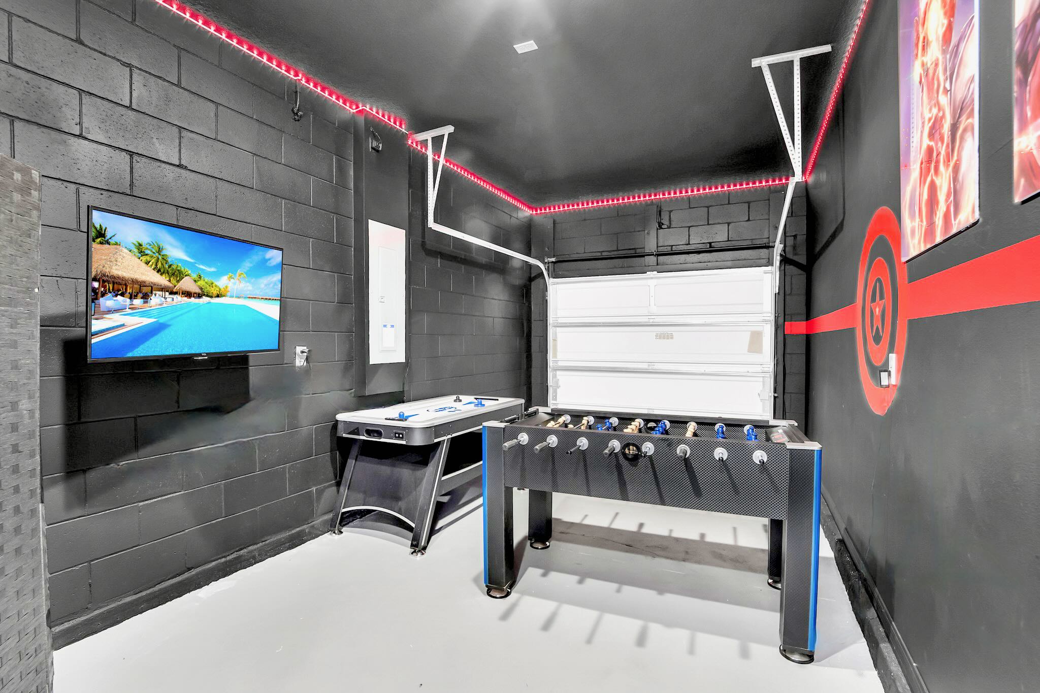 A converted garage game room features dark gray walls, red LED strip lighting, a foosball table, an air hockey table, and a wall-mounted TV, with the garage door visible in the background.