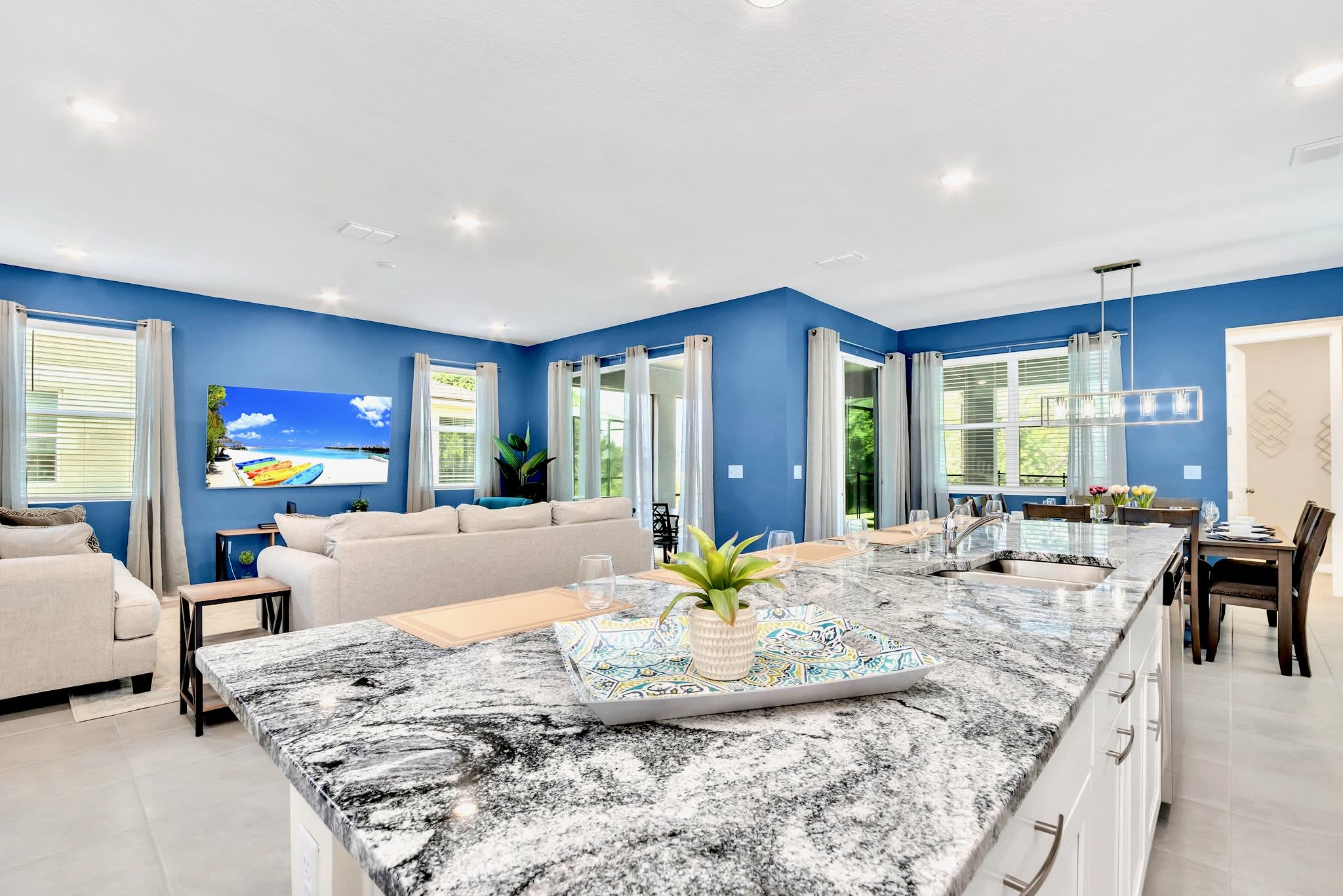 Modern kitchen island with a granite countertop, sink, and bar seating, offering an open view into the living room with blue walls and the dining area beyond. A decorative tray with a plant sits on the island.