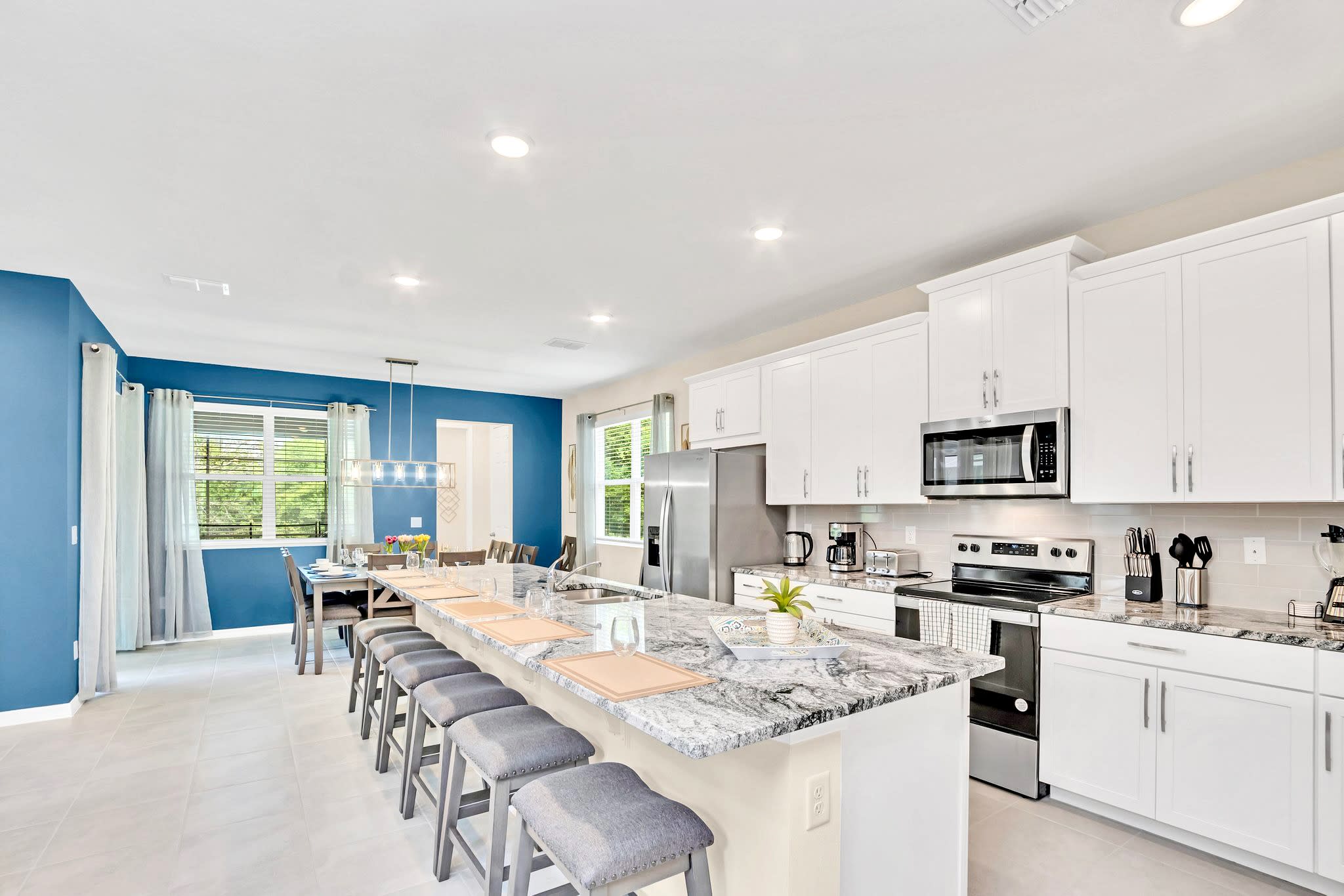 A modern kitchen features white shaker cabinets, stainless steel appliances, and a large granite island with seating for six, extending into a dining area with a long table and windows.