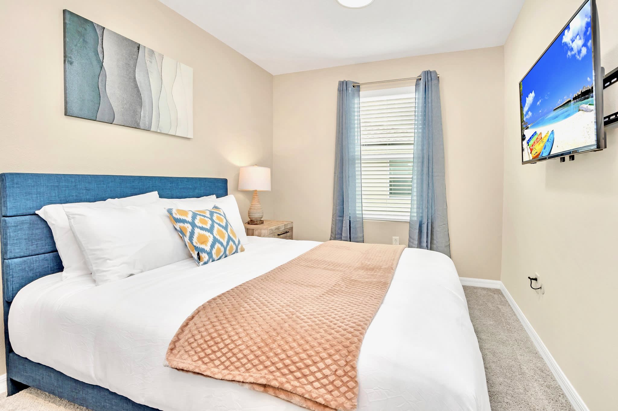 Cozy bedroom with a blue upholstered king-sized bed, white bedding, a patterned throw pillow, a wooden nightstand with a lamp, and a wall-mounted flat-screen TV.