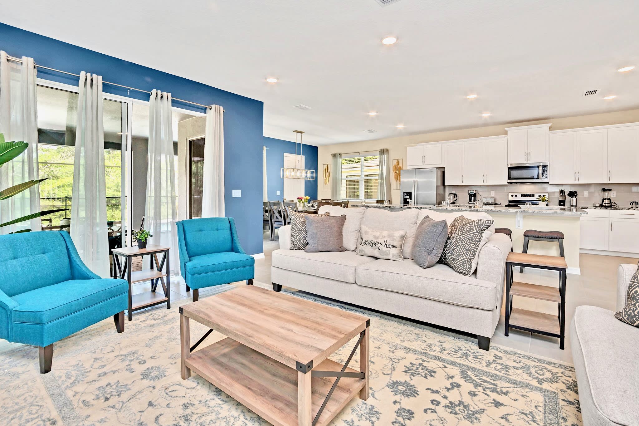 A bright open-concept living room features a light gray sofa, two turquoise armchairs, and a rustic wooden coffee table on a patterned rug, with a bold blue accent wall and a view into the kitchen and dining area.