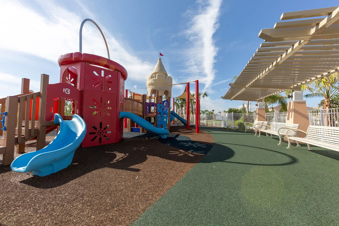A vibrant outdoor playground features a red and blue play structure with slides, a sand-colored castle-like tower, and multiple swings, set on a mix of rubber and wood chip ground cover under a bright sky. A white pergola with benches provides shade on the right.