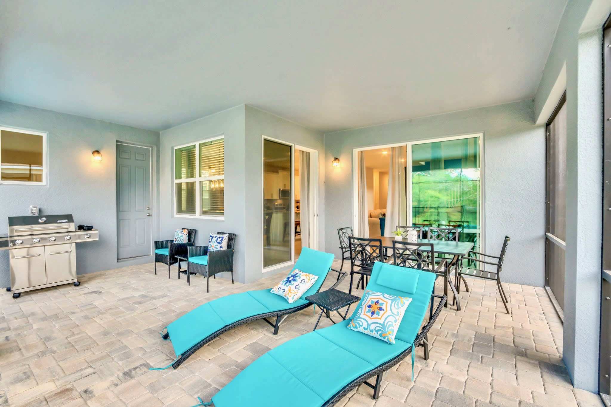 A spacious covered patio features two turquoise lounge chairs, an outdoor dining set, and a stainless steel BBQ grill, all on light pavers, with sliding glass doors leading into the house.