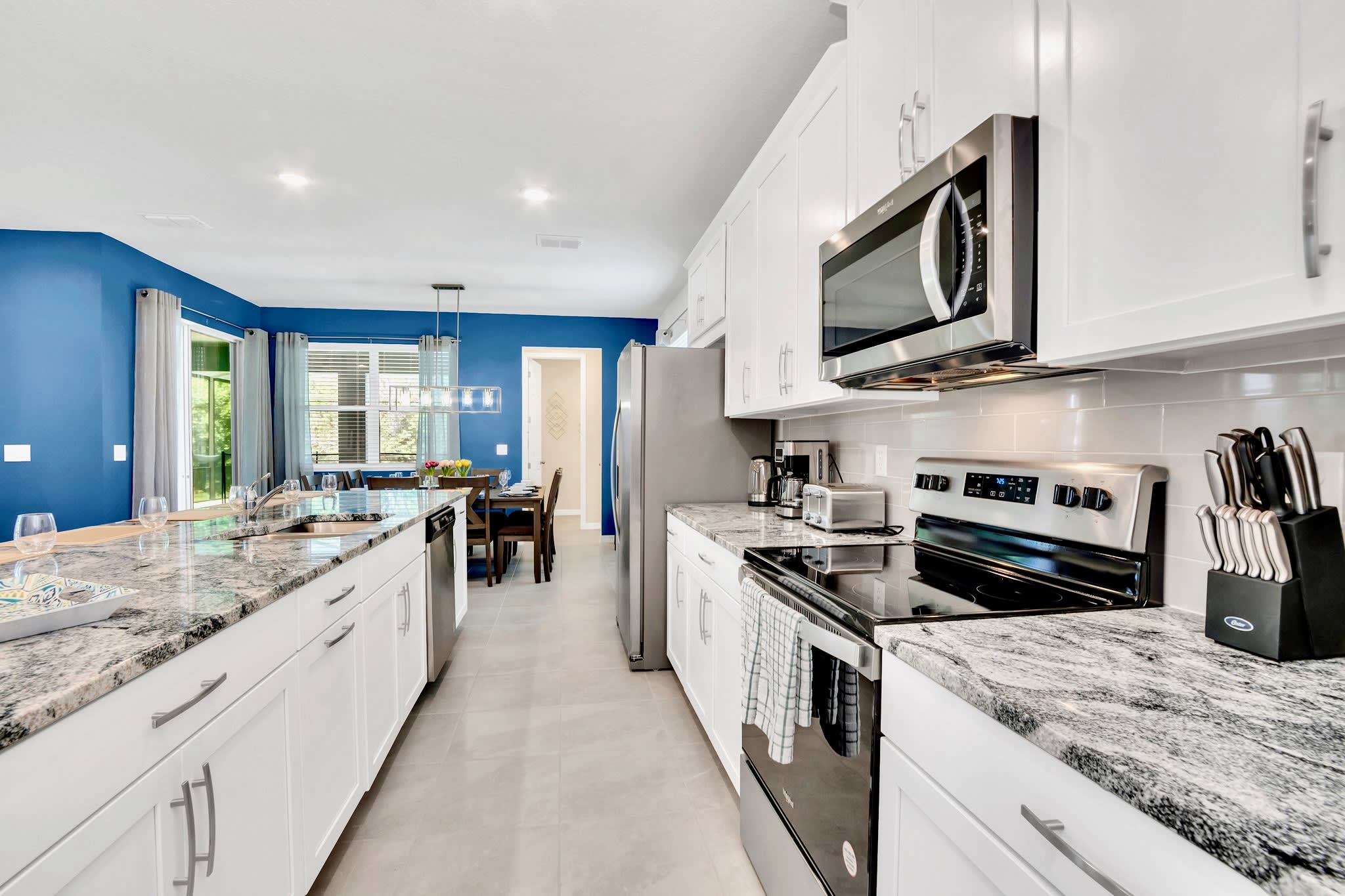 Galley-style kitchen with white cabinetry, stainless steel appliances including a refrigerator, oven, and microwave, and granite countertops. The dining area is visible in the background.