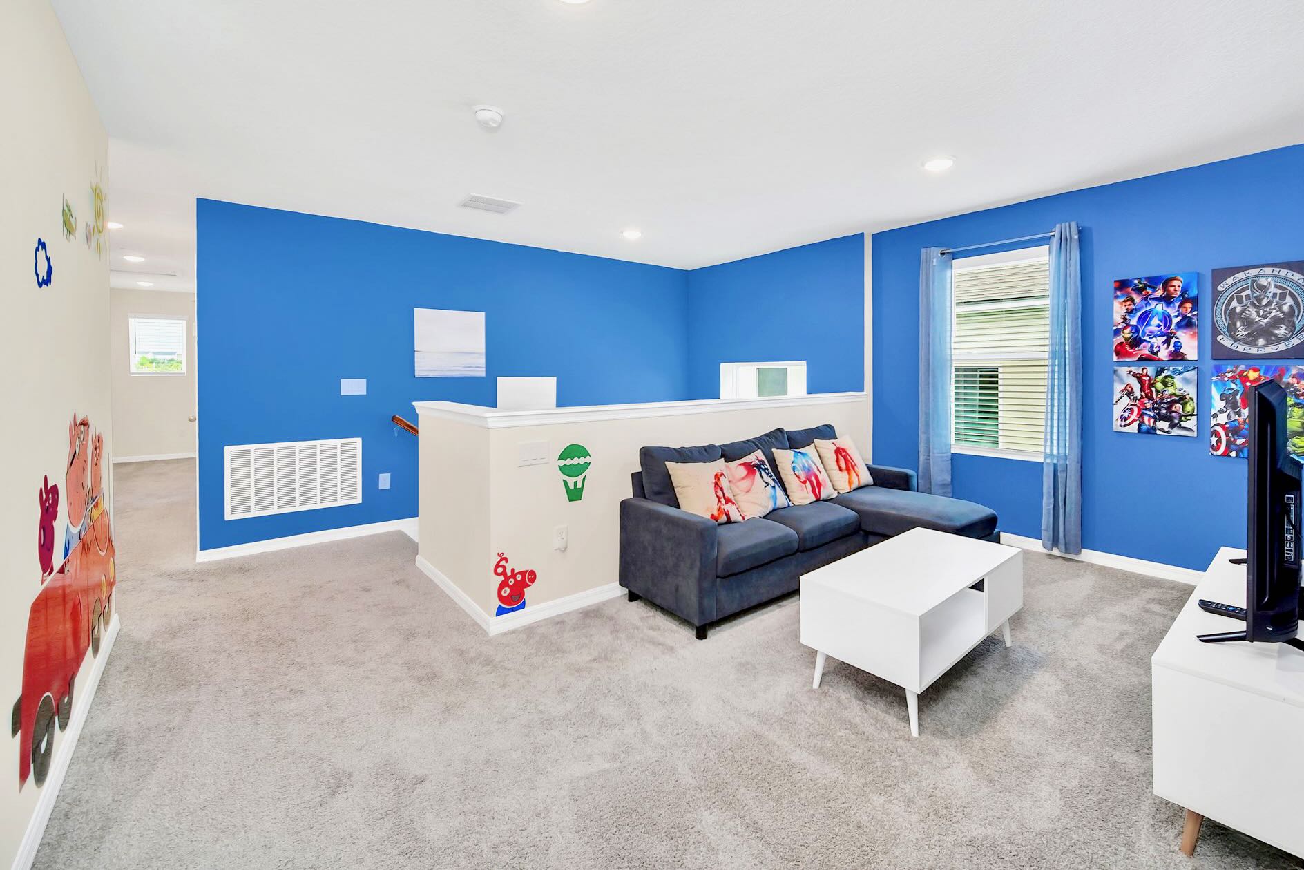Upstairs lounge area with vibrant blue walls, a dark blue sectional sofa, a white coffee table, and a TV stand. The space is decorated with superhero-themed wall decals and artwork.