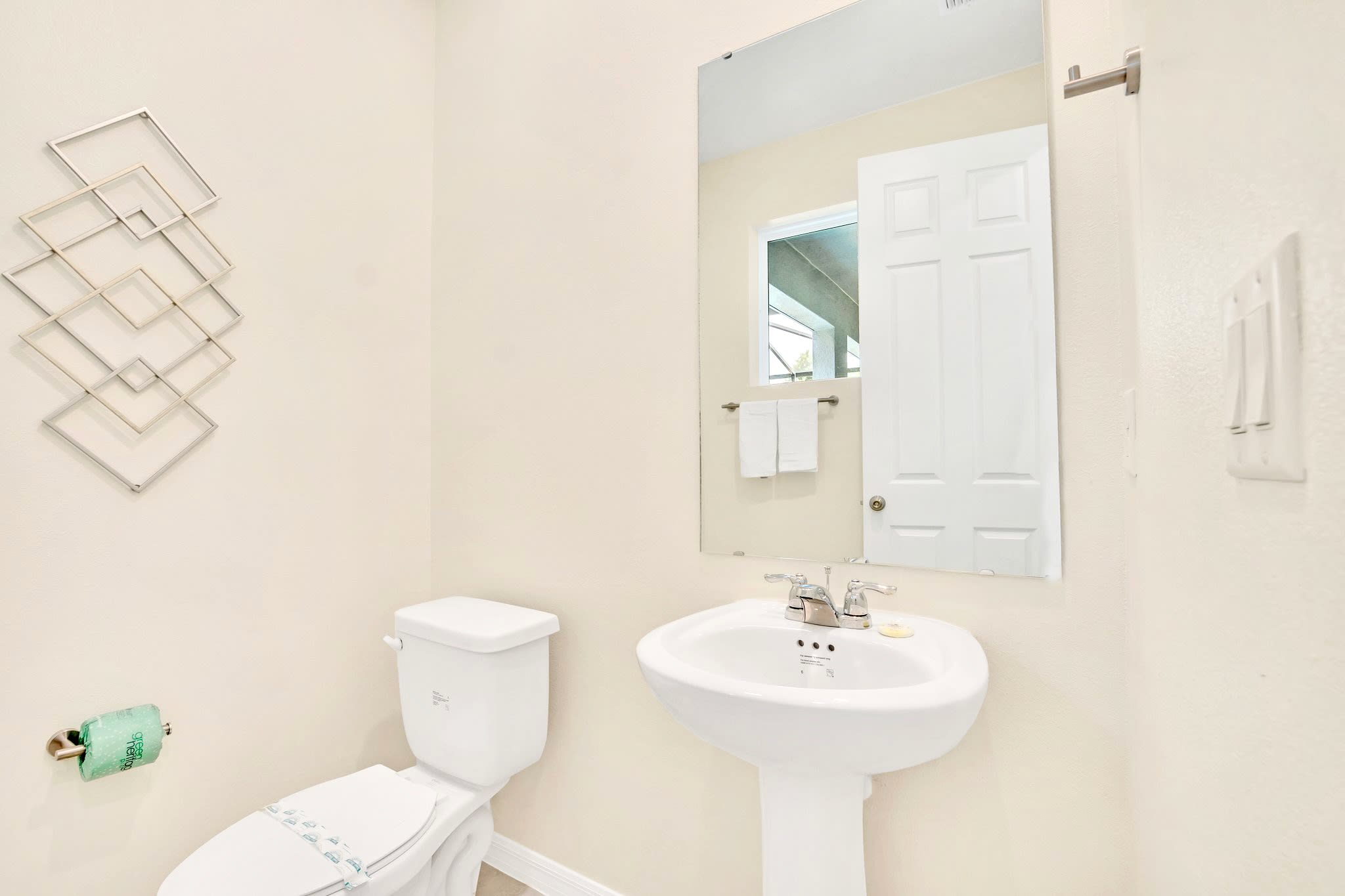 A bright, minimalist powder room features a white pedestal sink with a chrome faucet and a white toilet. A silver geometric wall art piece hangs above the toilet, and a large mirror reflects a white door and window.