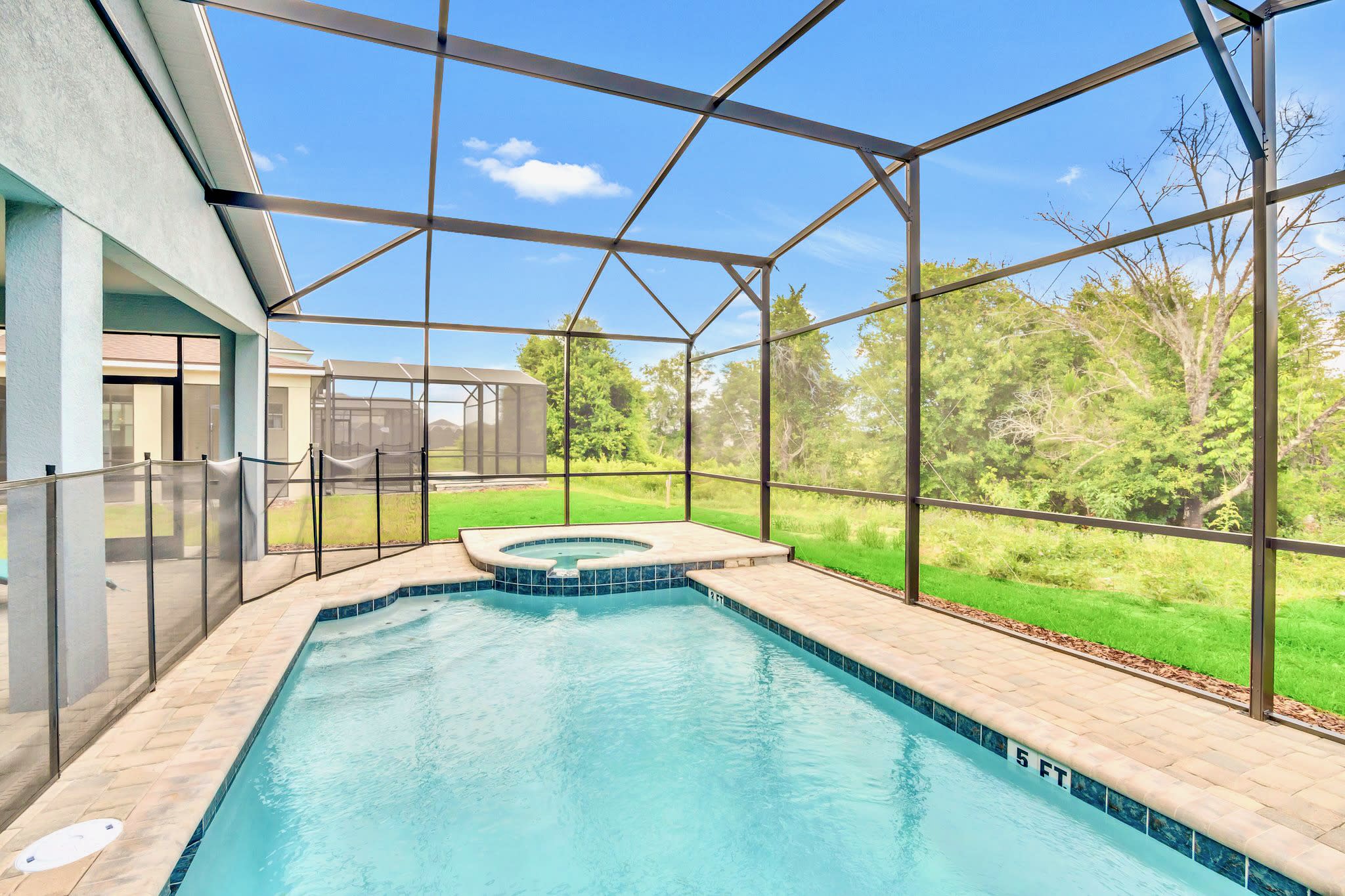A private screened-in pool area features a rectangular swimming pool with steps and a small hot tub, surrounded by light pavers and a safety fence, with a view of green grass and trees beyond the screen.