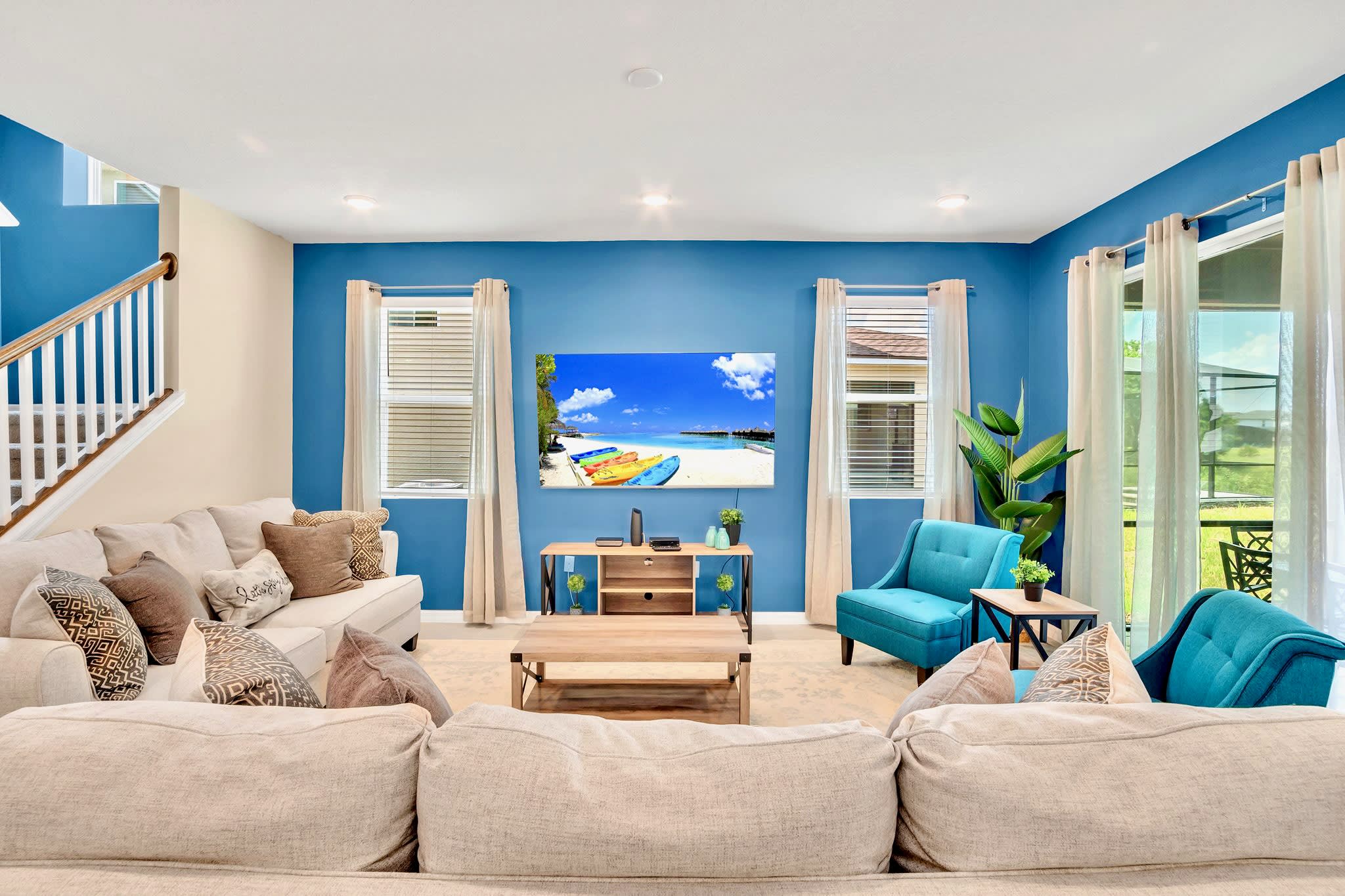 Bright living room featuring vibrant blue accent walls, two light-colored sofas, a large wall-mounted TV displaying a beach scene, and two windows with blinds and curtains.