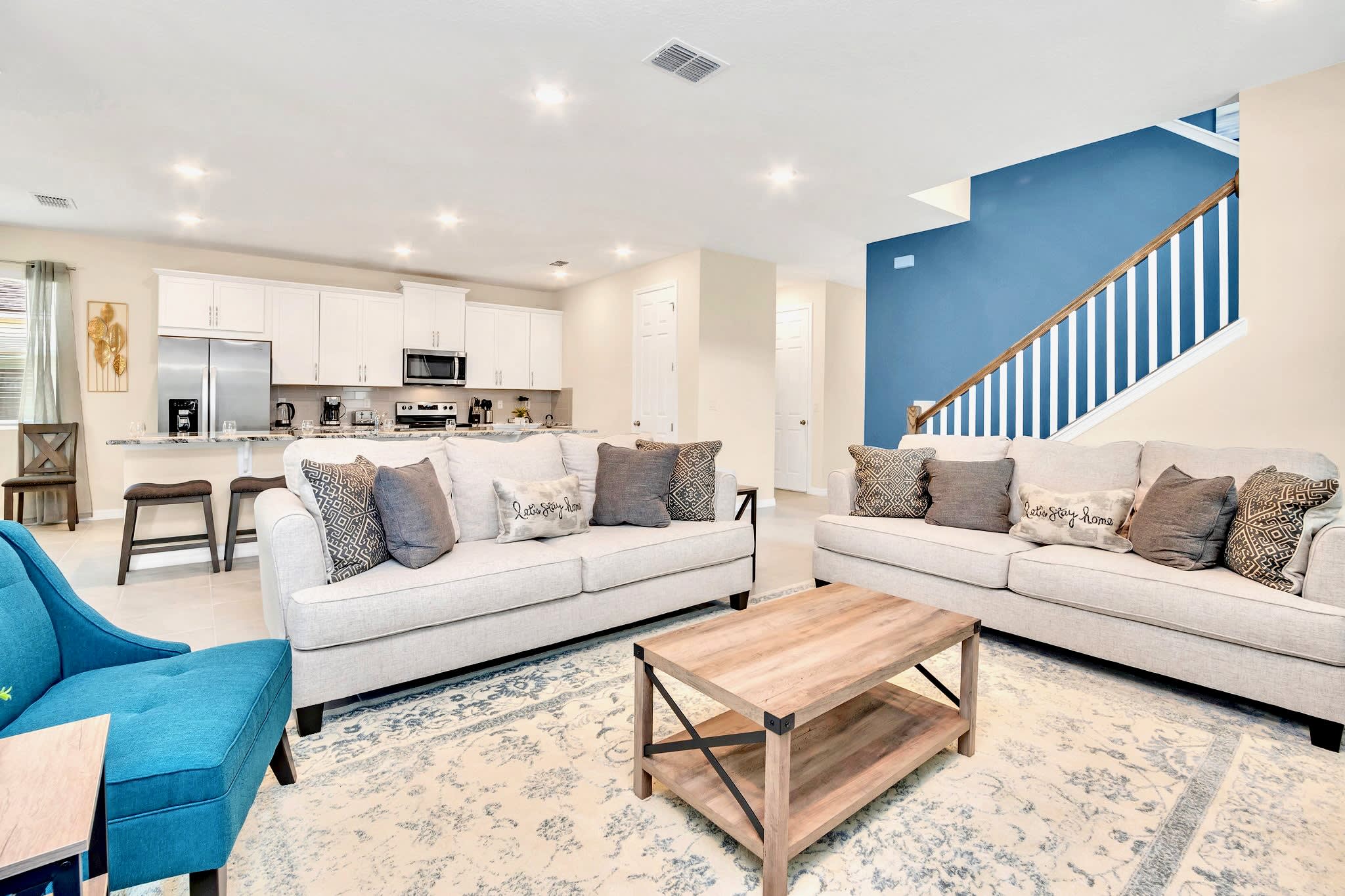 A bright open-concept living room features two light gray sofas and a turquoise armchair on a patterned rug, with a rustic wooden coffee table, and a staircase with a bold blue accent wall leading upstairs.