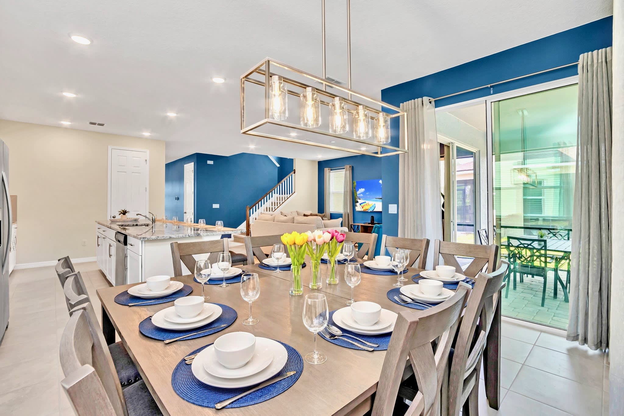 Spacious dining room with a large wooden table set for multiple guests, featuring blue placemats and white dinnerware, illuminated by a modern rectangular chandelier. The living room with blue walls is visible in the background.