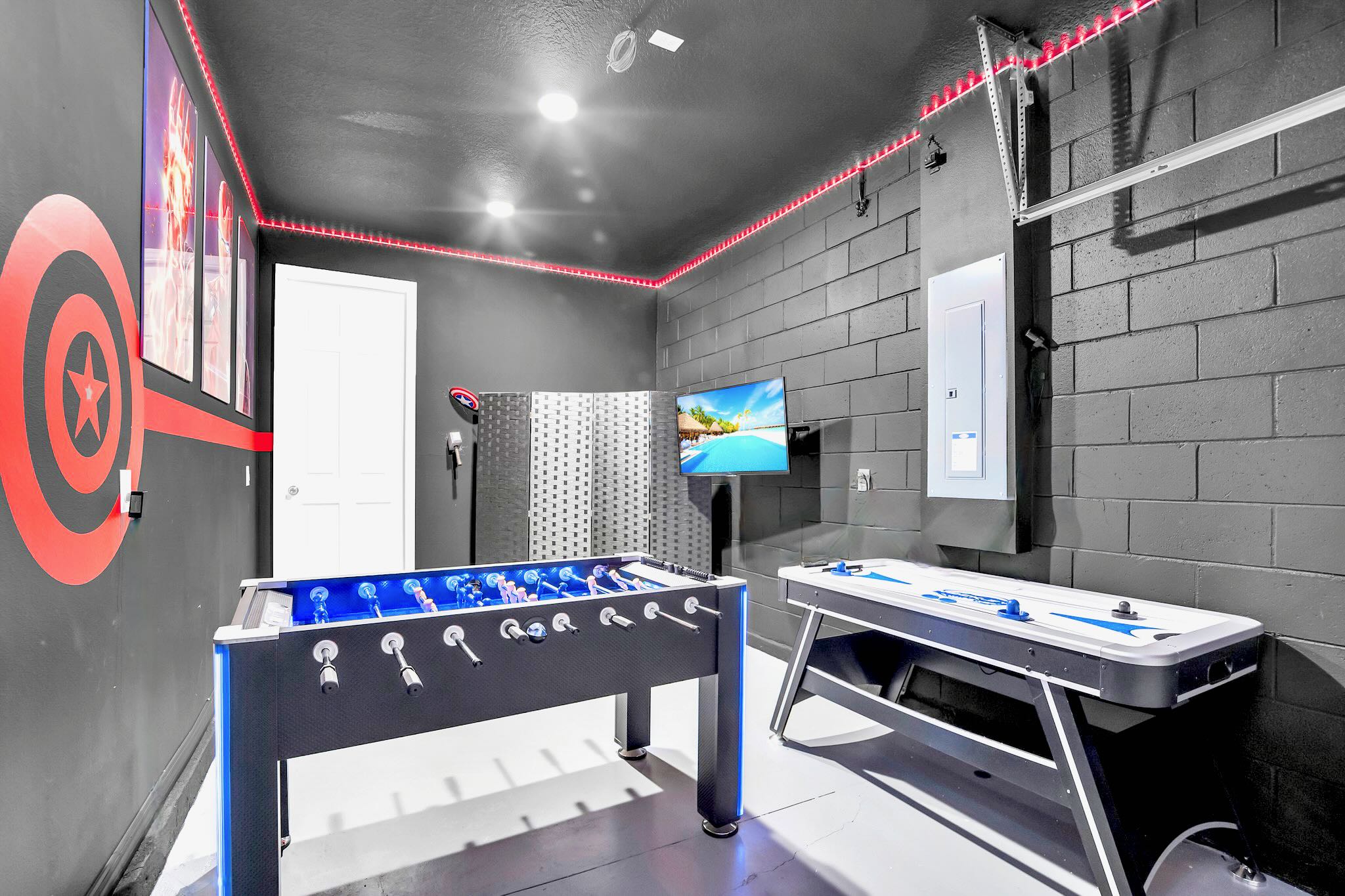 A vibrant game room with dark gray walls and ceiling features red LED strip lighting, a foosball table with blue accents, an air hockey table, and a wall-mounted TV, creating an energetic entertainment space.