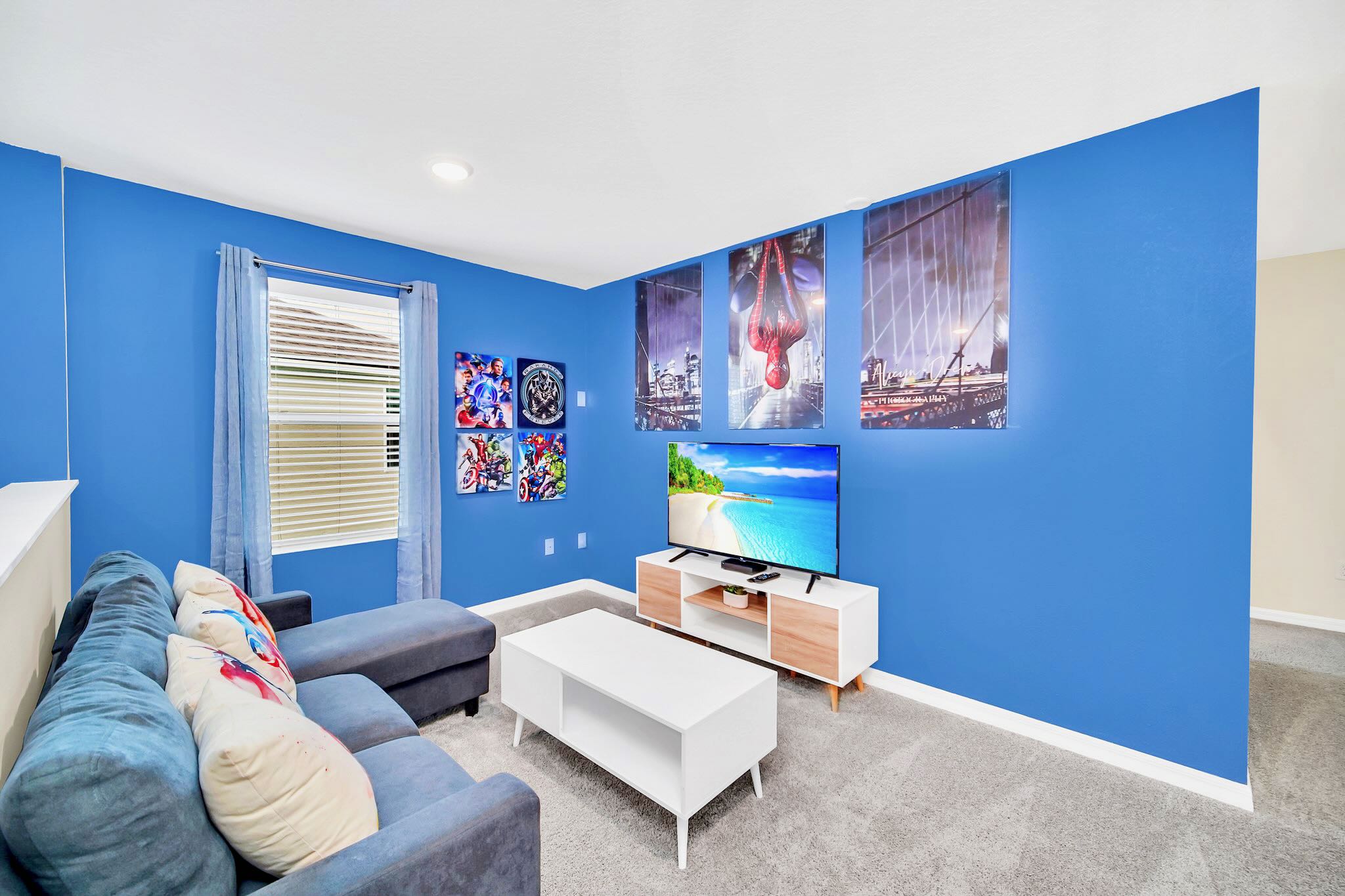 Another perspective of the upstairs media room, showcasing a wall-mounted TV on a modern white and wood console, surrounded by large superhero-themed posters. A dark blue sectional sofa is partially visible.