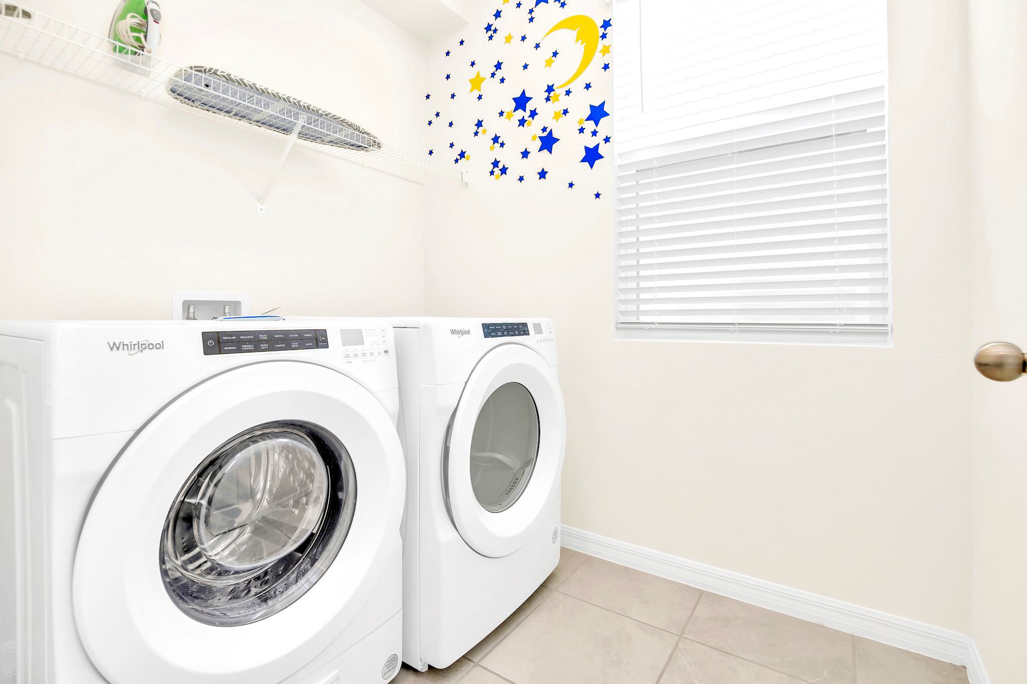 A modern laundry room with white Whirlpool front-loading washer and dryer units side-by-side. A wire shelf above holds an ironing board, and a playful blue and yellow star and moon decal decorates the wall.