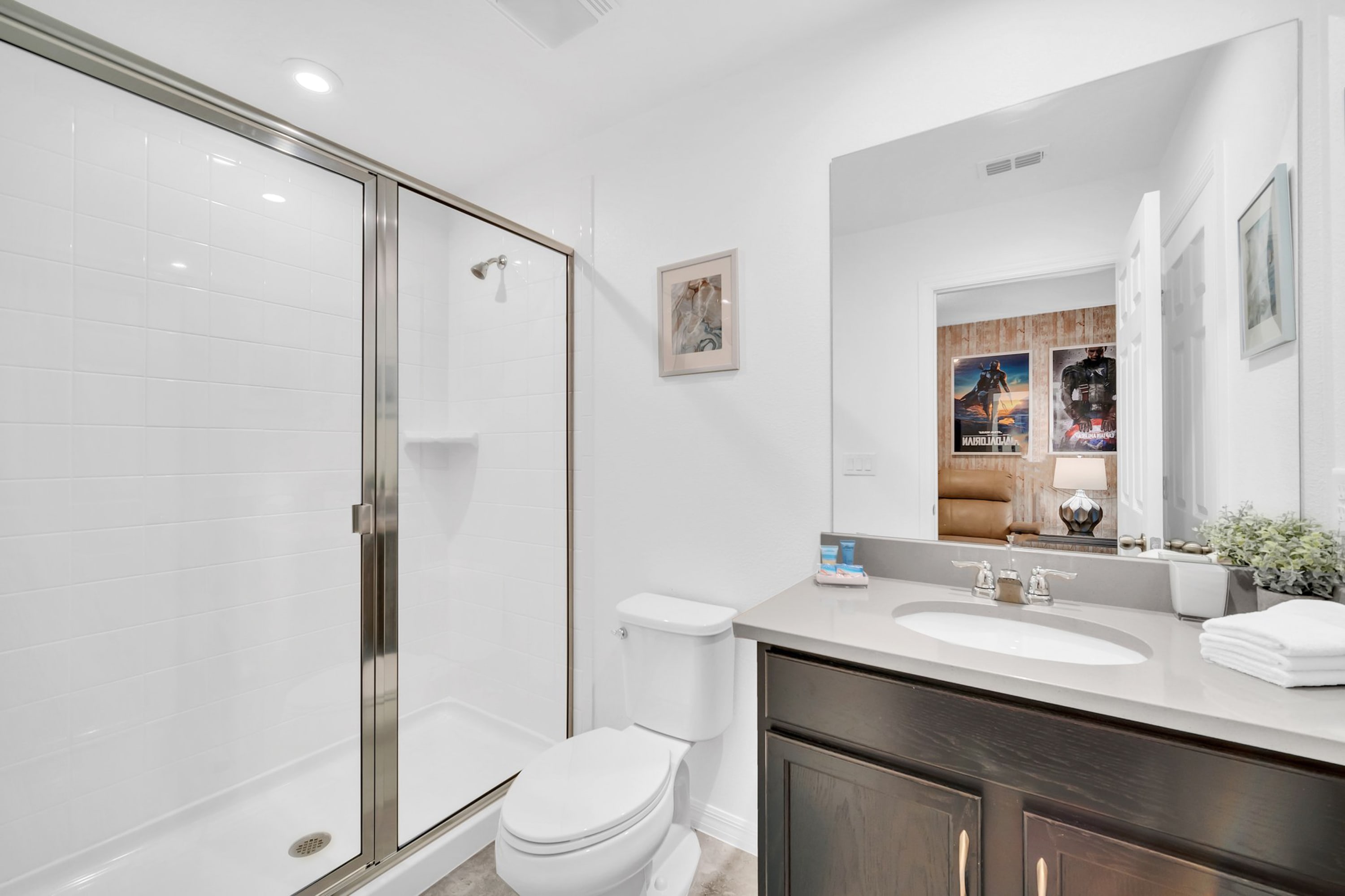 Modern bathroom with a glass-enclosed shower, white toilet, and a dark wood vanity with a light grey countertop and single sink. A framed picture hangs above the toilet.
