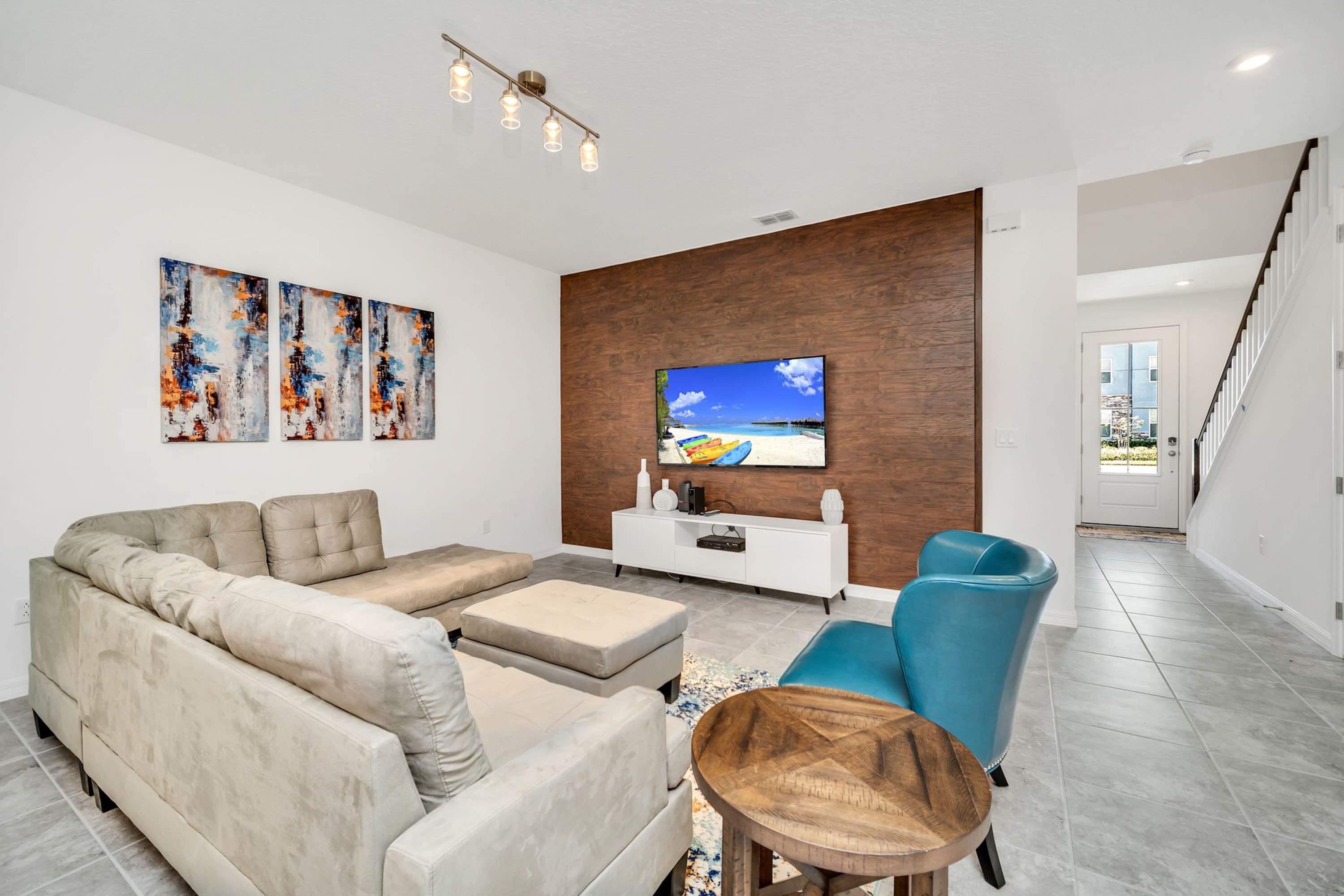 A modern living room features a large light beige sectional sofa facing a wall with a wooden accent panel and a mounted flat-screen TV. A white media console sits below the TV, and a blue accent chair and patterned rug complete the space.