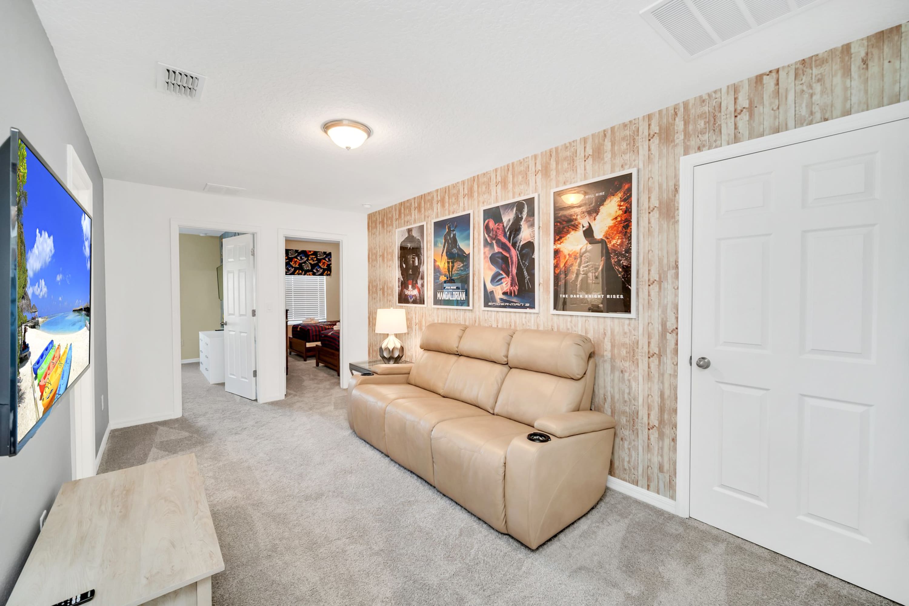 Cozy media room featuring a light brown reclining sofa, a large wall-mounted TV, and movie posters adorning a light wood-paneled accent wall, with soft gray carpeted flooring.