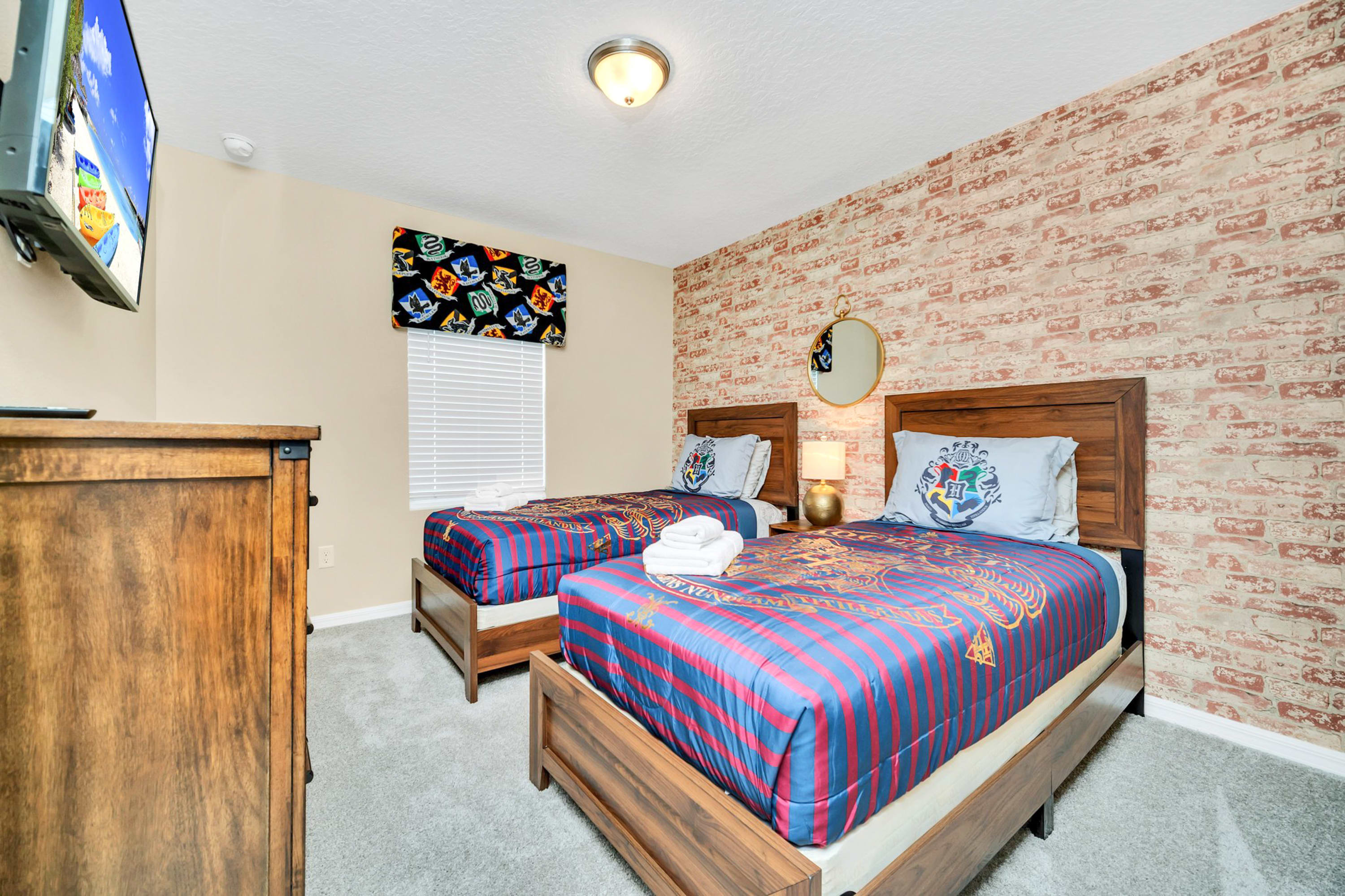 Themed bedroom with two twin beds featuring Harry Potter bedding, a brick-patterned accent wall, and a small window with blinds. A wooden dresser and wall-mounted TV are visible.
