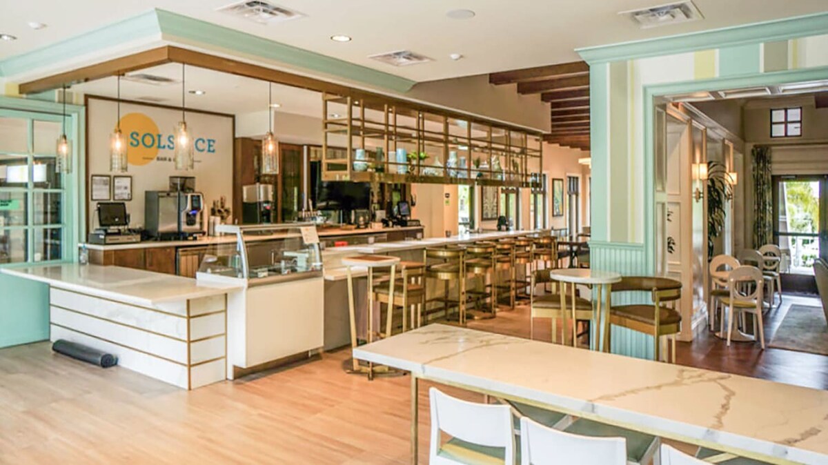 A bright, modern bar and cafe area features a long counter with a display case, gold-accented shelving, and numerous bar stools. The space has light wood floors, light green and white walls, and a sign for "SOLSTICE BAR & CAFE".