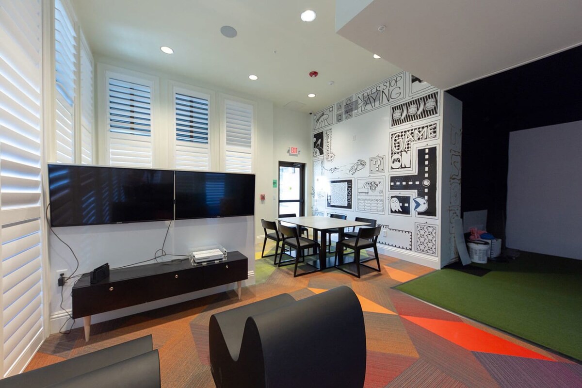 A modern indoor game room or lounge area featuring two large wall-mounted TVs, a black console, a square dining table with chairs, and a colorful geometric carpet transitioning to a green turf area.