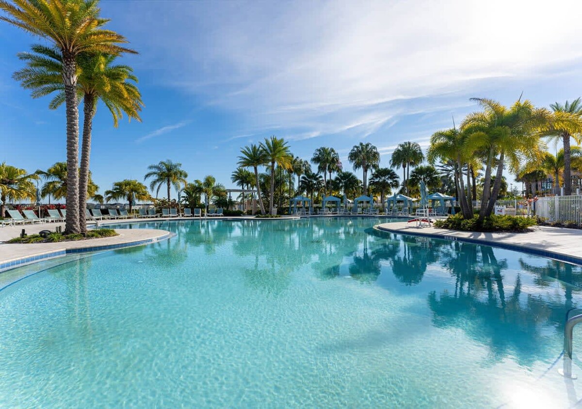 A large, irregularly shaped communal swimming pool with clear blue water, surrounded by numerous palm trees and lounge chairs under a bright blue sky with scattered clouds.