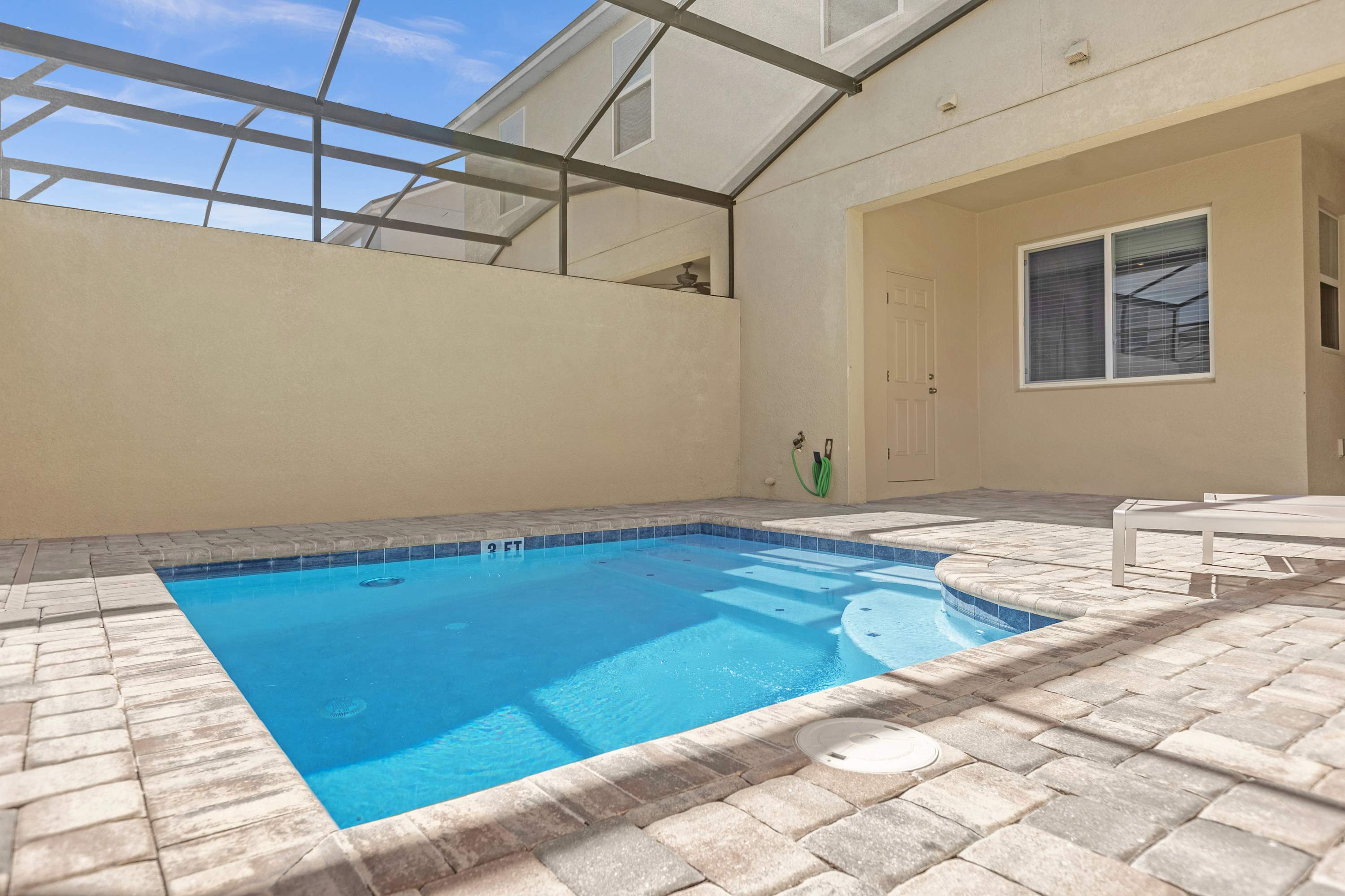 A screened-in private pool area features a small rectangular pool with a shallow end, surrounded by light-colored pavers. The patio is covered, and a door leads into the house, with a hose and outdoor equipment visible.