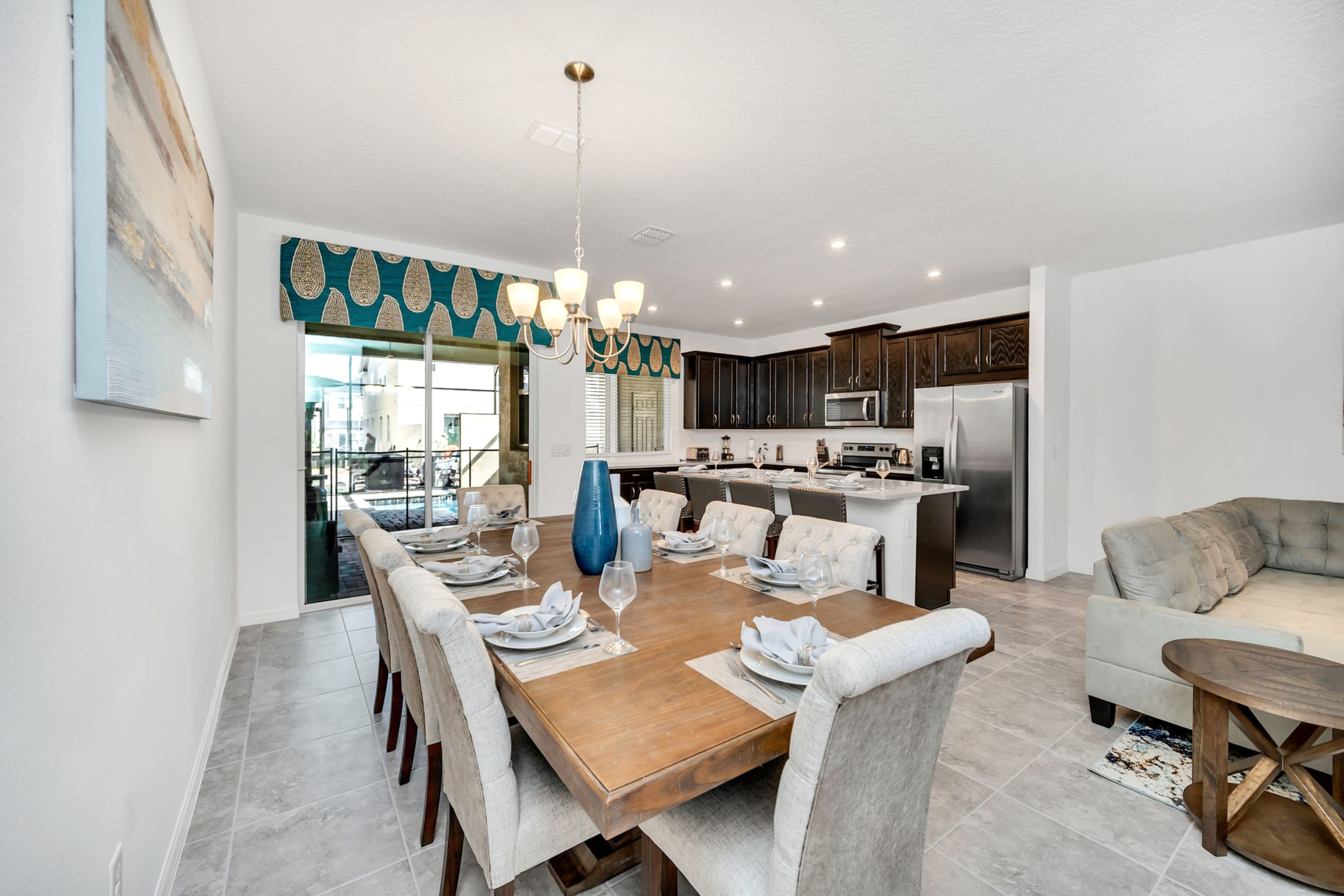 Spacious dining room featuring a long wooden table set for multiple guests, surrounded by upholstered chairs, with a view of the kitchen and living area through a sliding glass door.
