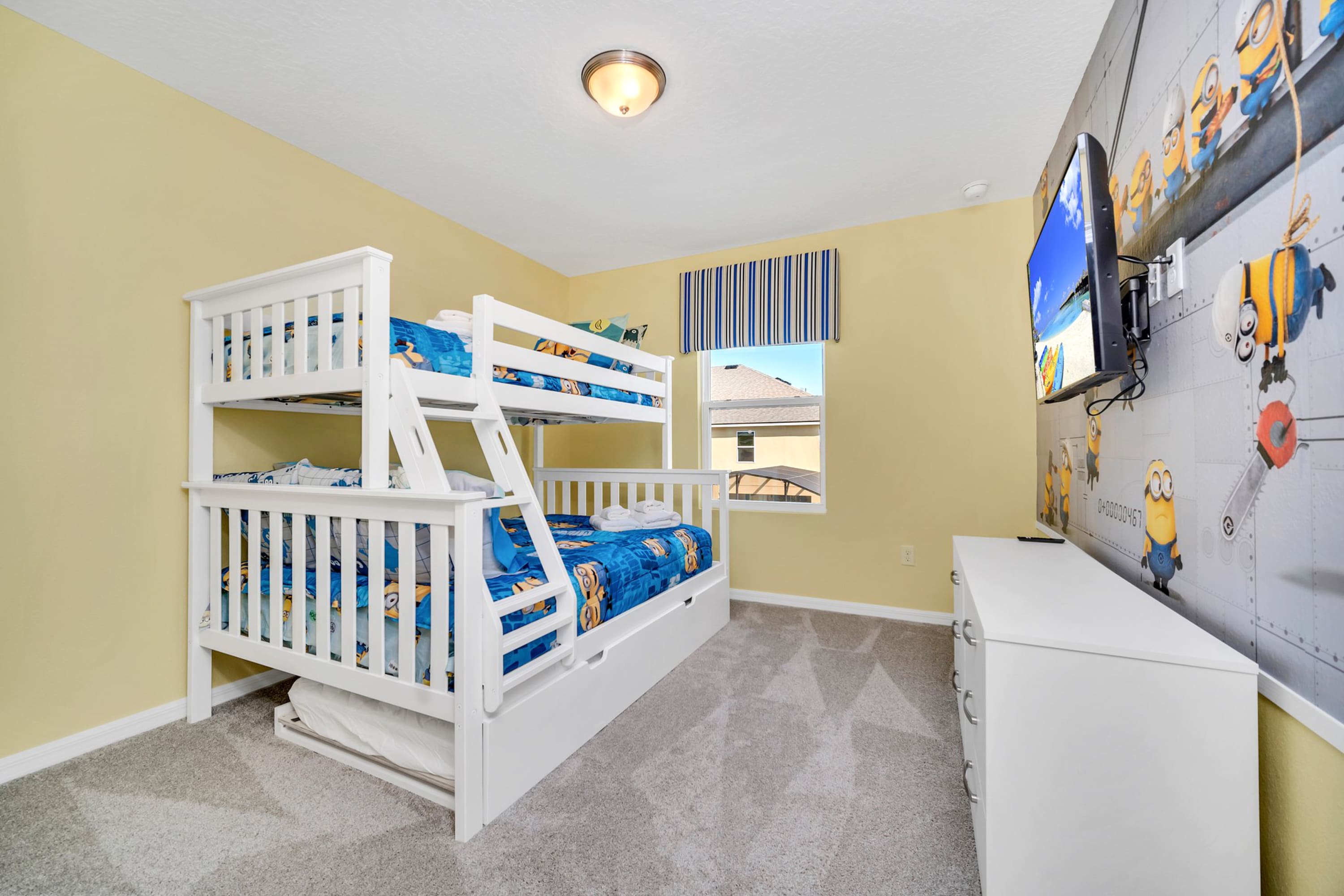 Playful kids' bedroom with a white bunk bed and trundle, Minions-themed bedding, and a large Minions wall mural. A white dresser and wall-mounted TV complete the space.