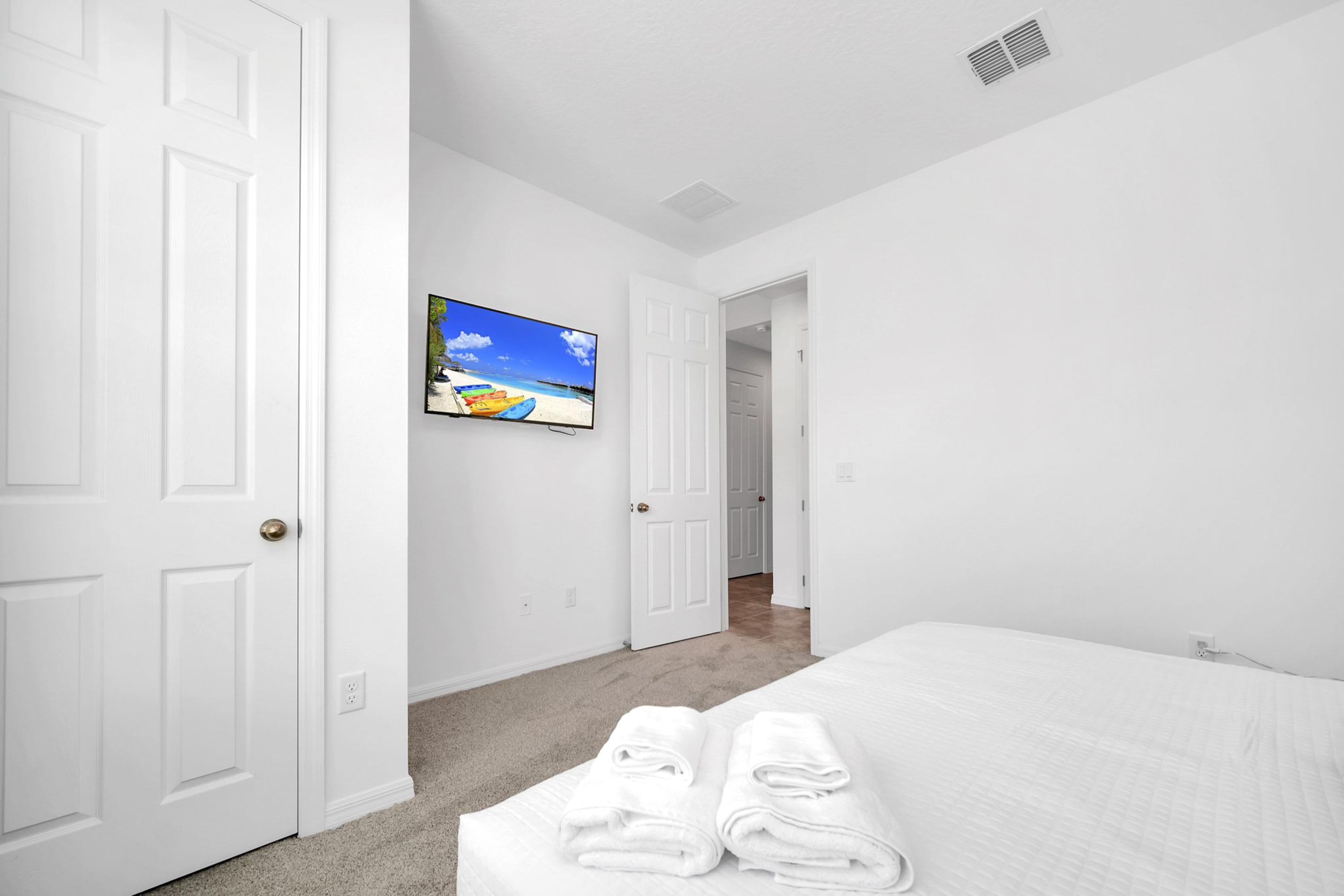 Bright bedroom with white walls and carpeted floor, featuring a queen-sized bed with white bedding and two folded white towels, and a wall-mounted TV.