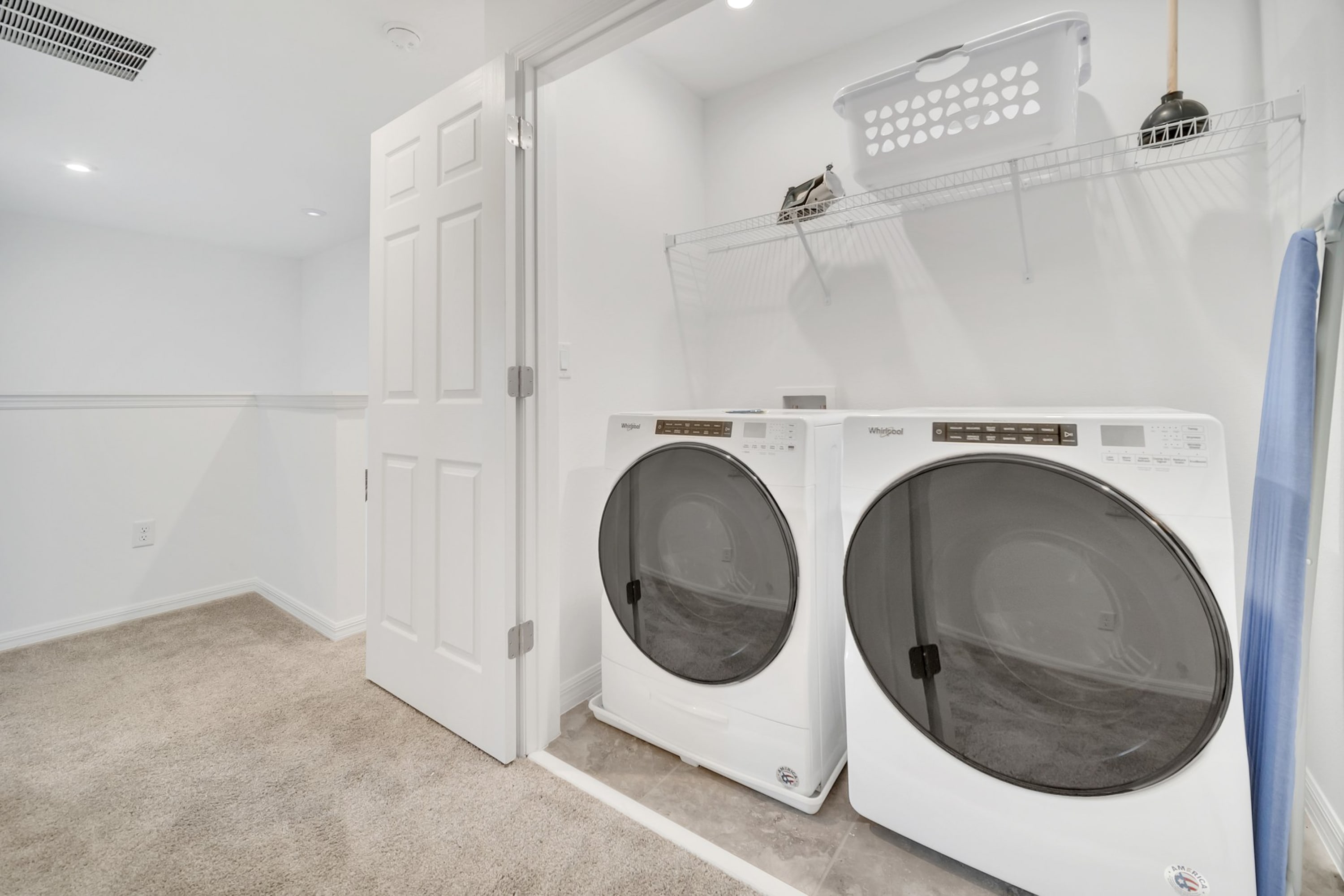 Dedicated laundry area with side-by-side white front-loading washer and dryer units, a wire shelf above, and a laundry basket. The room has carpeted flooring and white walls.