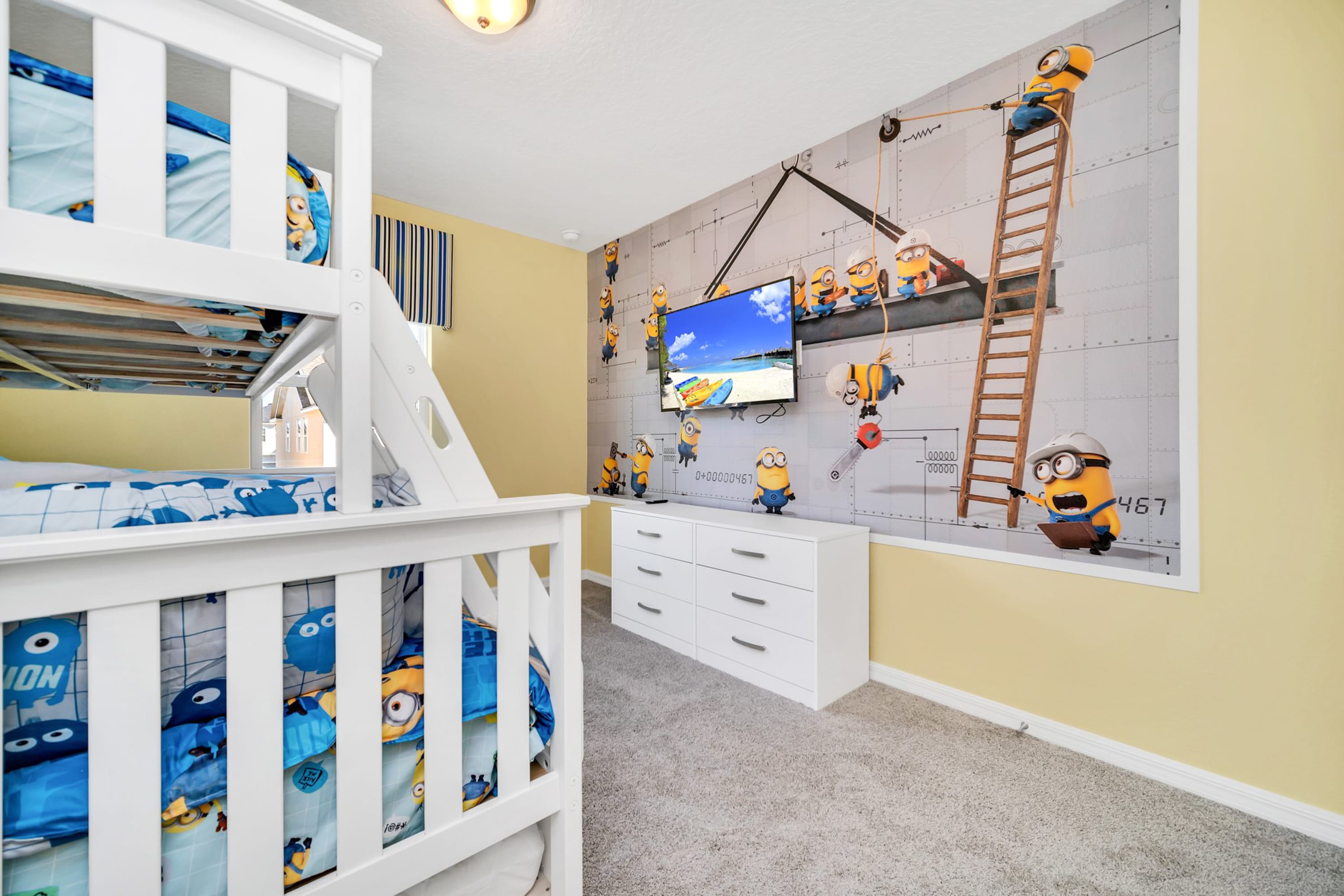 Close-up view of the Minions-themed bedroom, highlighting the large wall mural with various Minions characters and a white dresser below a wall-mounted TV.
