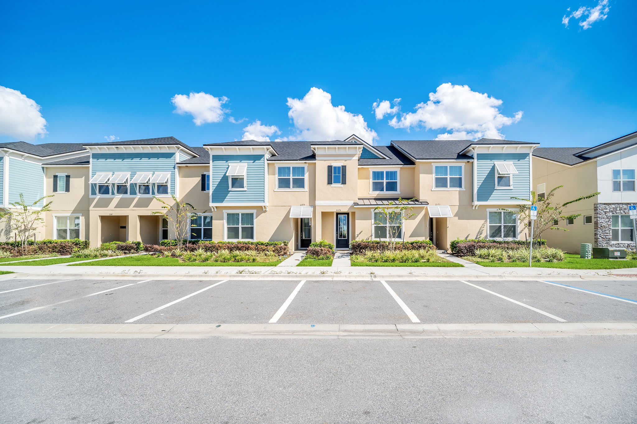 Front exterior view of a modern townhouse complex with light blue and beige facades, multiple windows, and neatly landscaped front yards. A paved parking lot is in the foreground under a clear blue sky.
