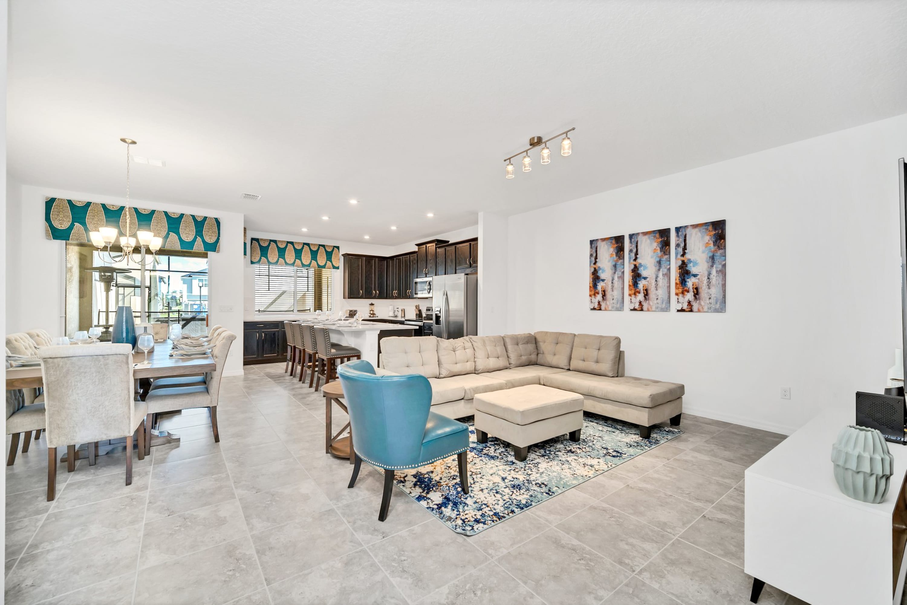 An open-concept living space features a light beige sectional sofa, a blue accent chair, and a dining area with a long wooden table. The kitchen with dark cabinetry and a large island is visible in the background, all set on light gray tiled floors.