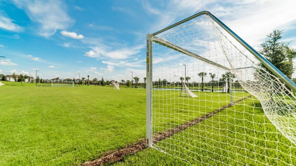 A wide-angle view of a well-maintained green soccer field with multiple white goalposts, stretching under a bright blue sky with fluffy white clouds, surrounded by distant trees and residential buildings.