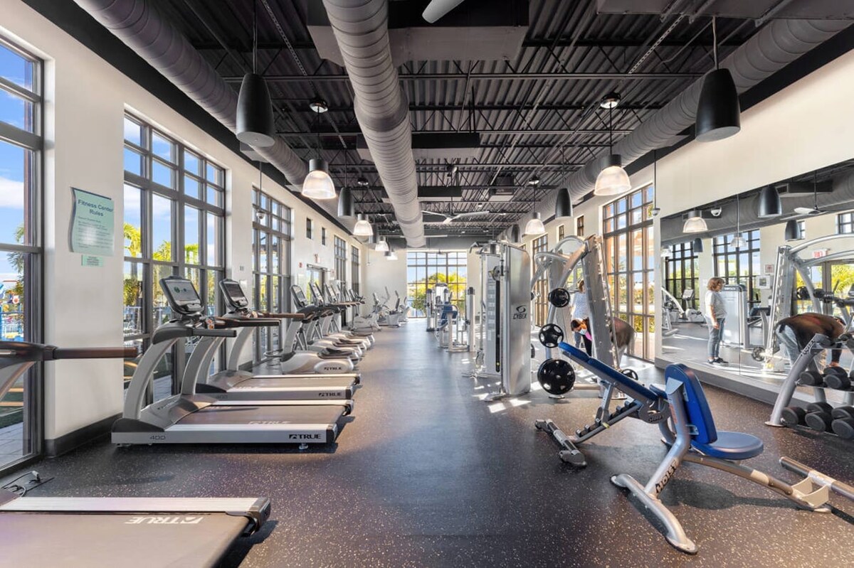 A large, well-equipped fitness center features rows of treadmills, weight machines, and free weights, all set on a dark speckled floor. Floor-to-ceiling windows provide ample natural light and views, while the ceiling has exposed dark ductwork and industrial lighting.
