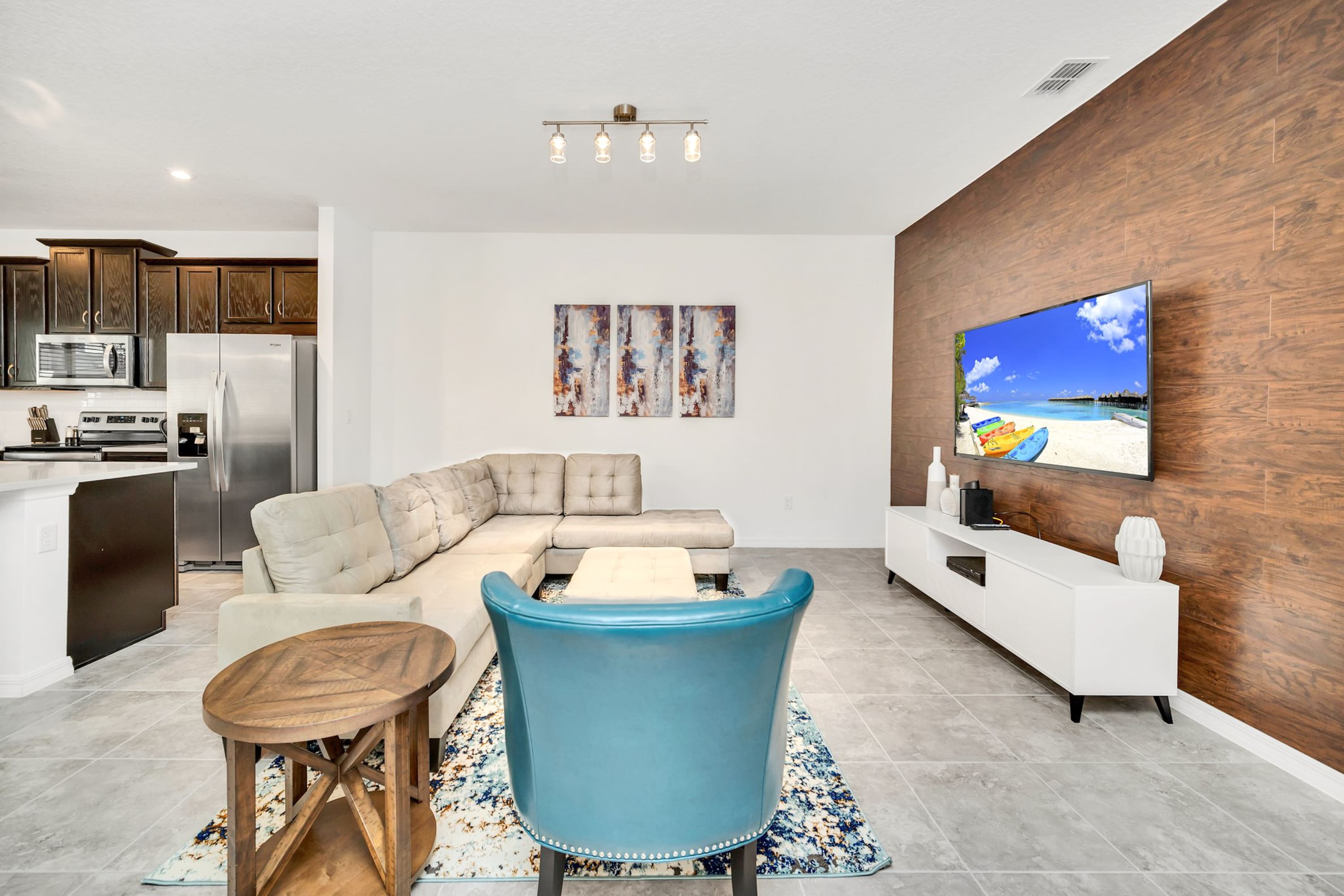 Modern living room with a large L-shaped sectional sofa, a blue accent chair, and a TV mounted on a wood-paneled accent wall, featuring light gray tiled floors and a view into the kitchen.