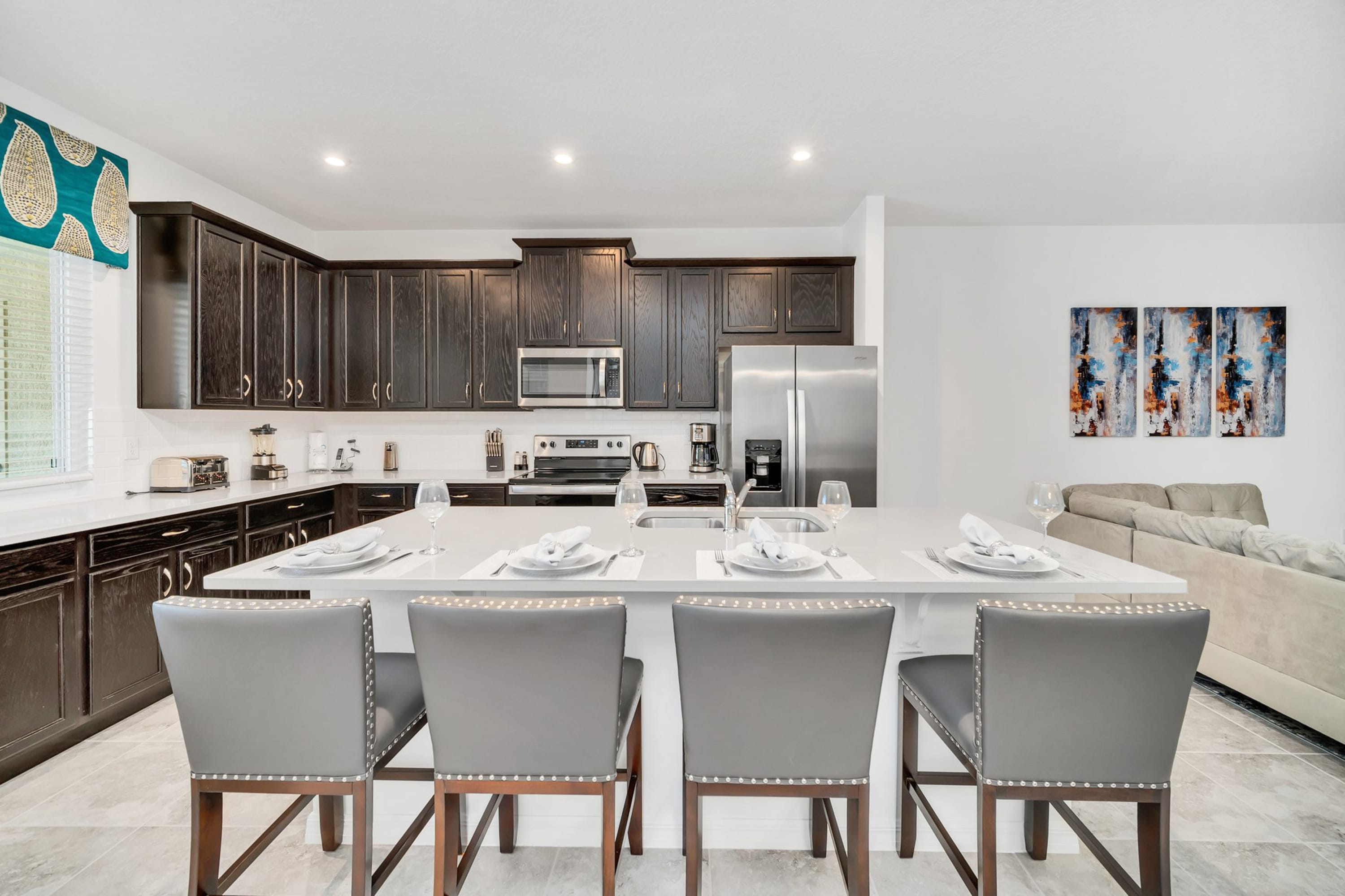 A modern kitchen features dark wood cabinetry, white countertops, and stainless steel appliances, including a refrigerator, microwave, and oven. A large island with four gray upholstered bar stools is set with place settings, all on light gray tiled floors.