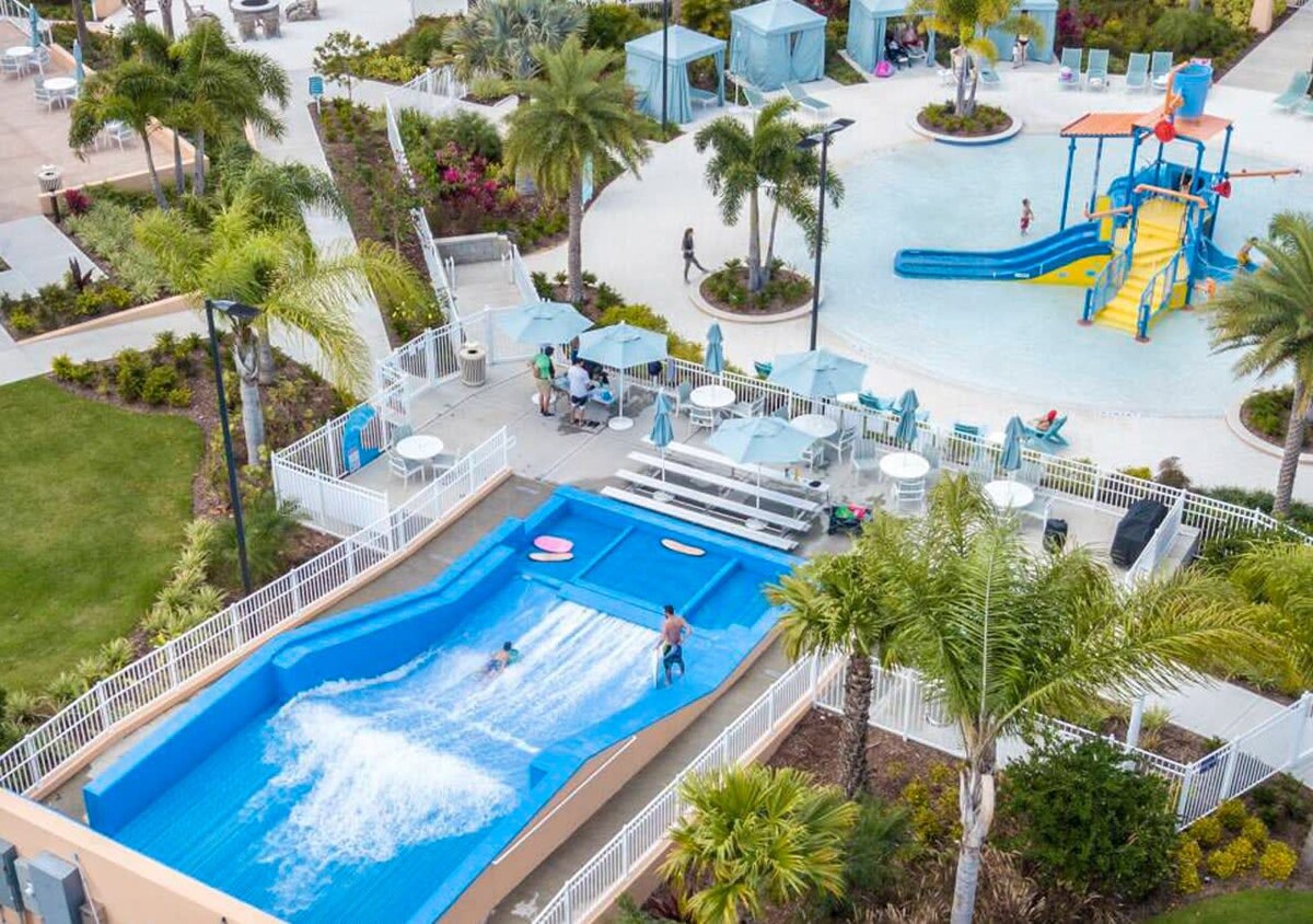 An aerial view of a vibrant water park area featuring a blue surf simulator with a person riding a wave, a shallow splash pad with a small water slide, and various seating areas with umbrellas.