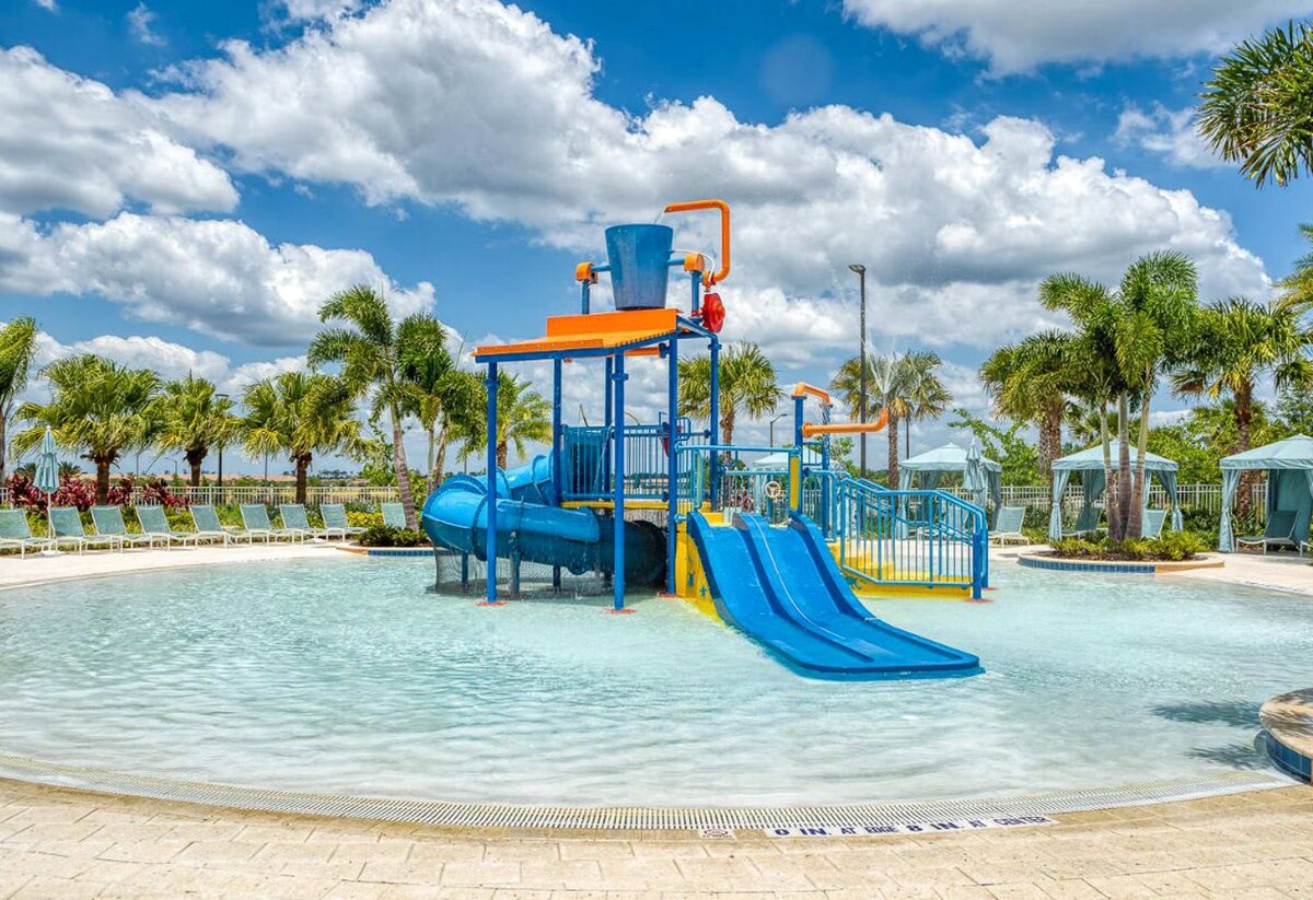 A shallow, zero-entry pool designed for children, featuring a blue and orange water play structure with slides, a bucket dump, and various spray elements under a bright, cloudy sky.