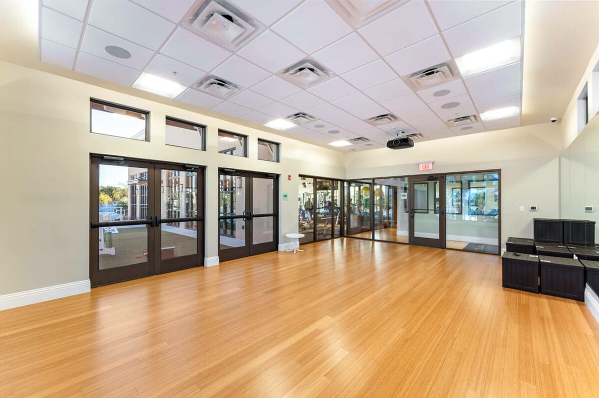 A spacious, empty multi-purpose room features light wood flooring, a drop ceiling with recessed lighting, and large mirrors along one wall. Multiple glass doors lead to other areas, suggesting it's part of a larger facility.