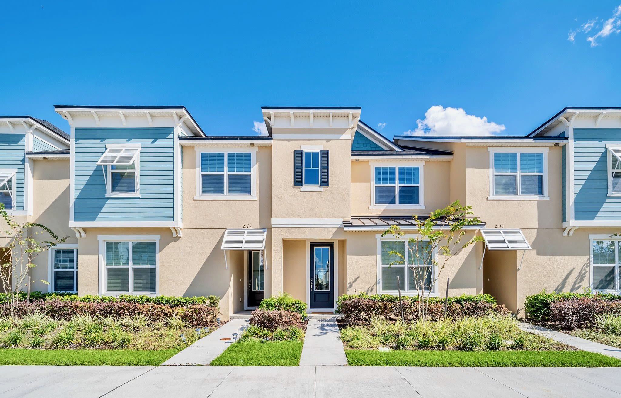 The front exterior of a modern townhouse features a light beige facade with blue and white accents on the upper level. Multiple windows with white trim and a dark front door are visible, surrounded by manicured landscaping and a concrete walkway.