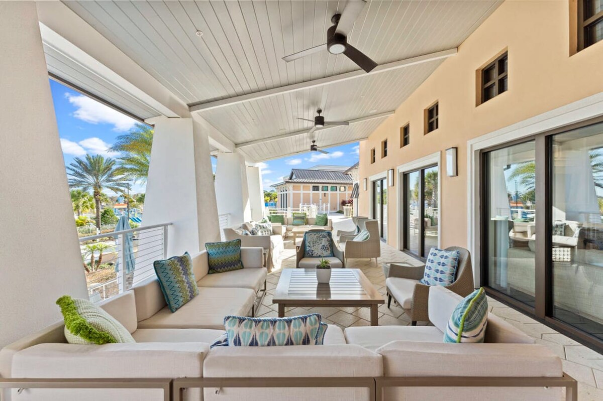 A spacious covered outdoor patio features light-colored sectional sofas with colorful accent pillows, ceiling fans, and views of palm trees and a resort-style area. The space is furnished with modern outdoor furniture and has a light, airy feel.