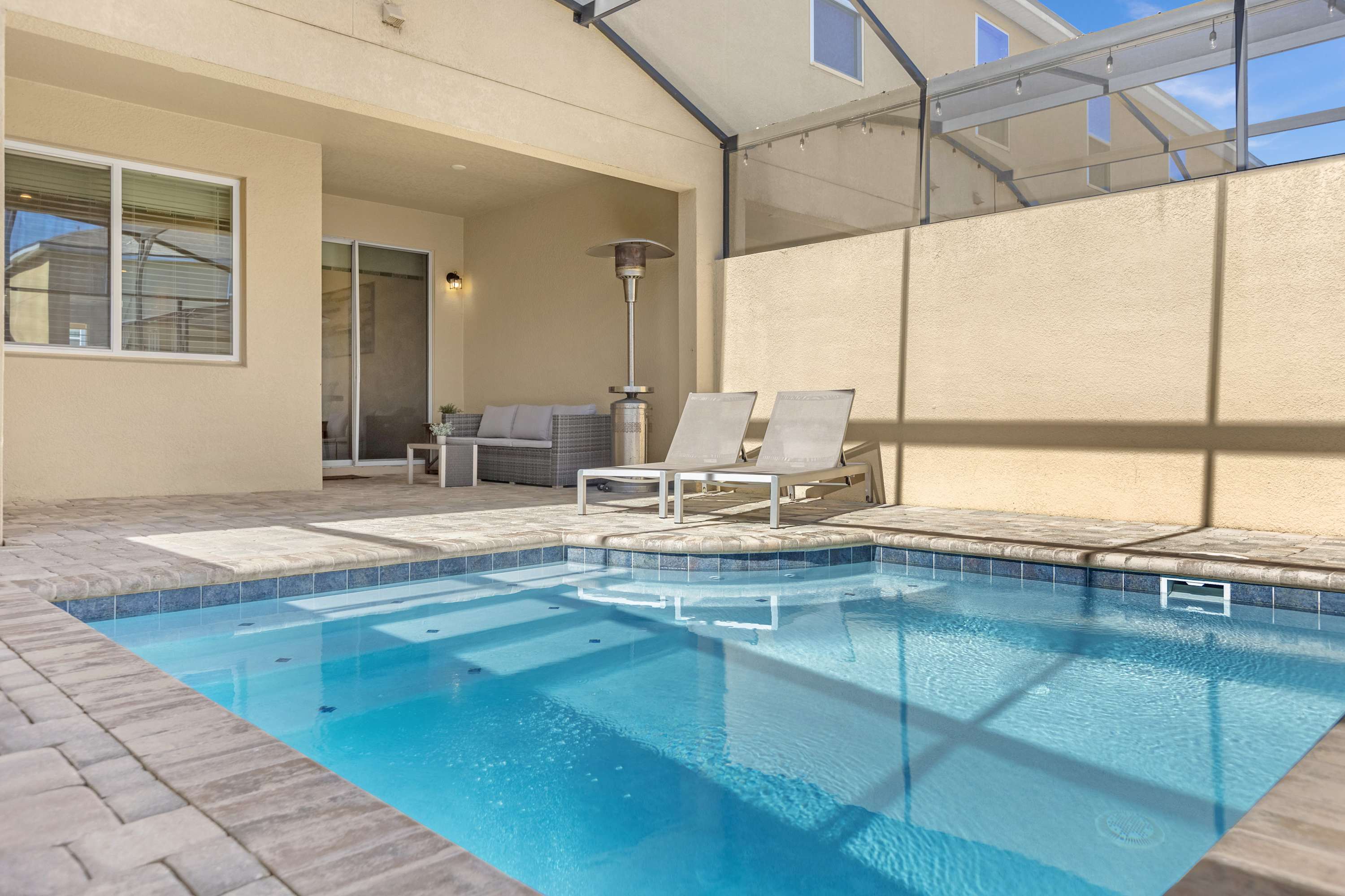 A screened-in private pool area features a small rectangular pool with steps, surrounded by light-colored pavers. A patio heater and two lounge chairs are visible on the covered patio, with a light beige stucco wall enclosing the space.