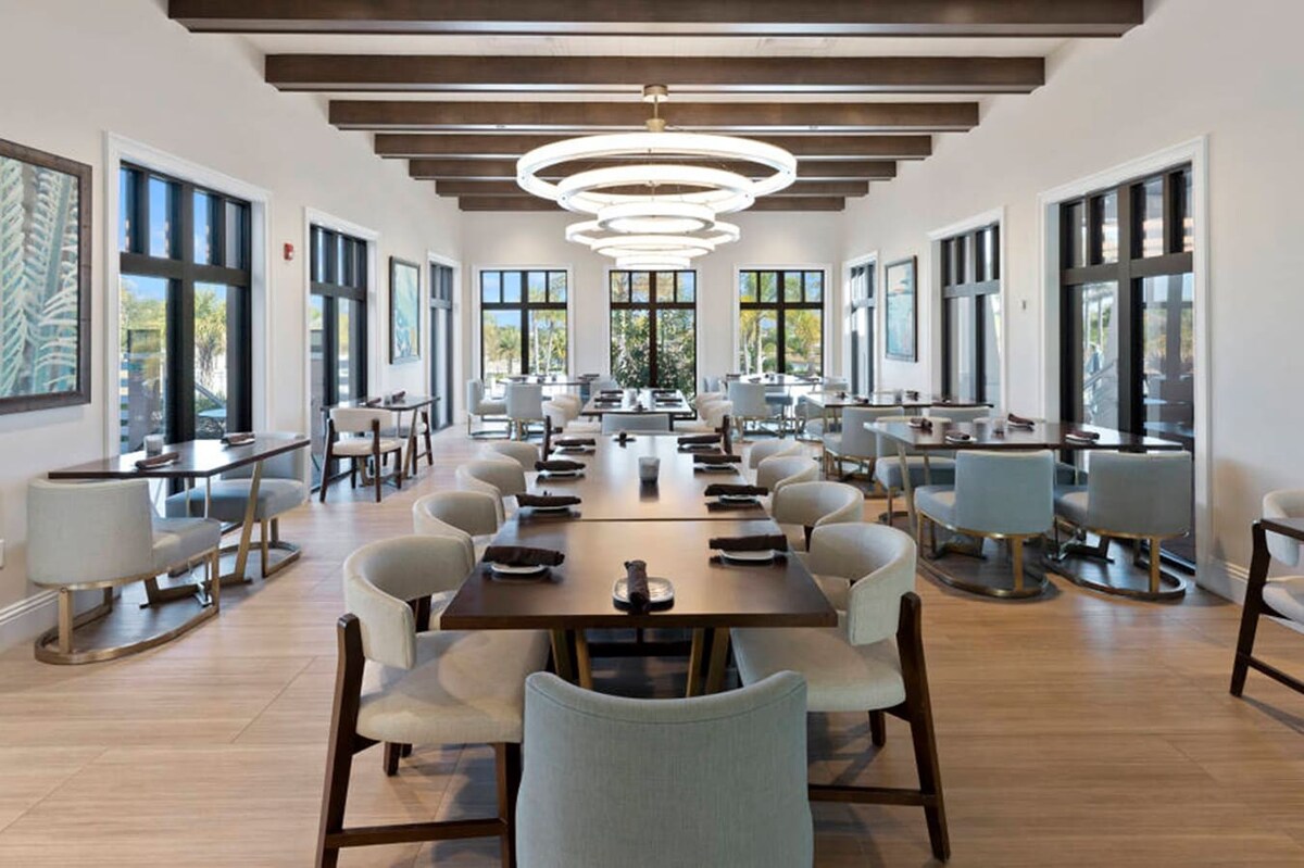 A sophisticated dining room features multiple long dark wood tables surrounded by light-colored upholstered chairs, illuminated by a series of modern circular chandeliers. Large windows line the walls, offering views of the outdoors, and the ceiling has dark exposed beams.
