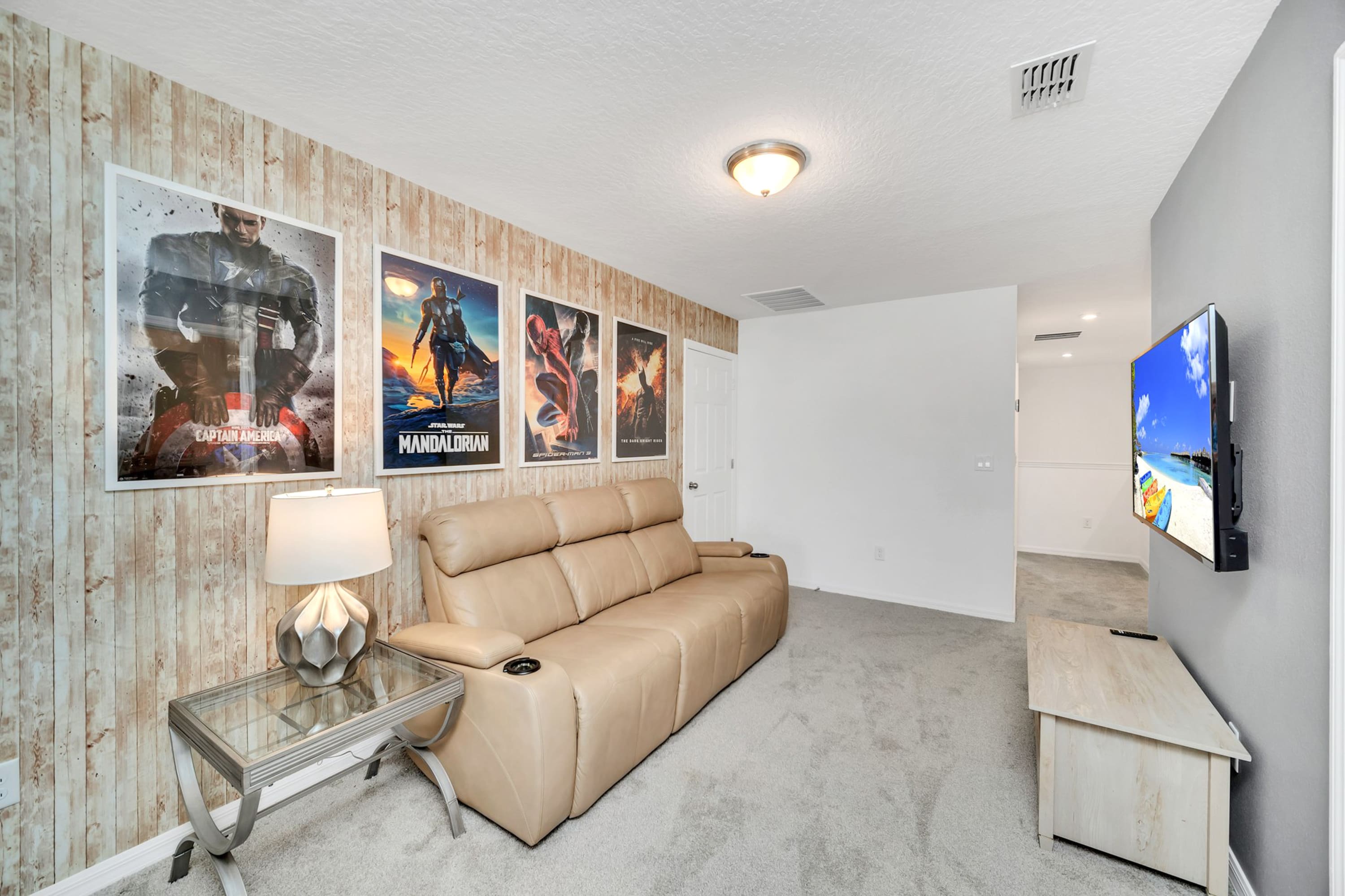 Alternate view of the media room, highlighting the light brown reclining sofa, glass-top side table with a lamp, and a series of movie posters on the wood-paneled wall.