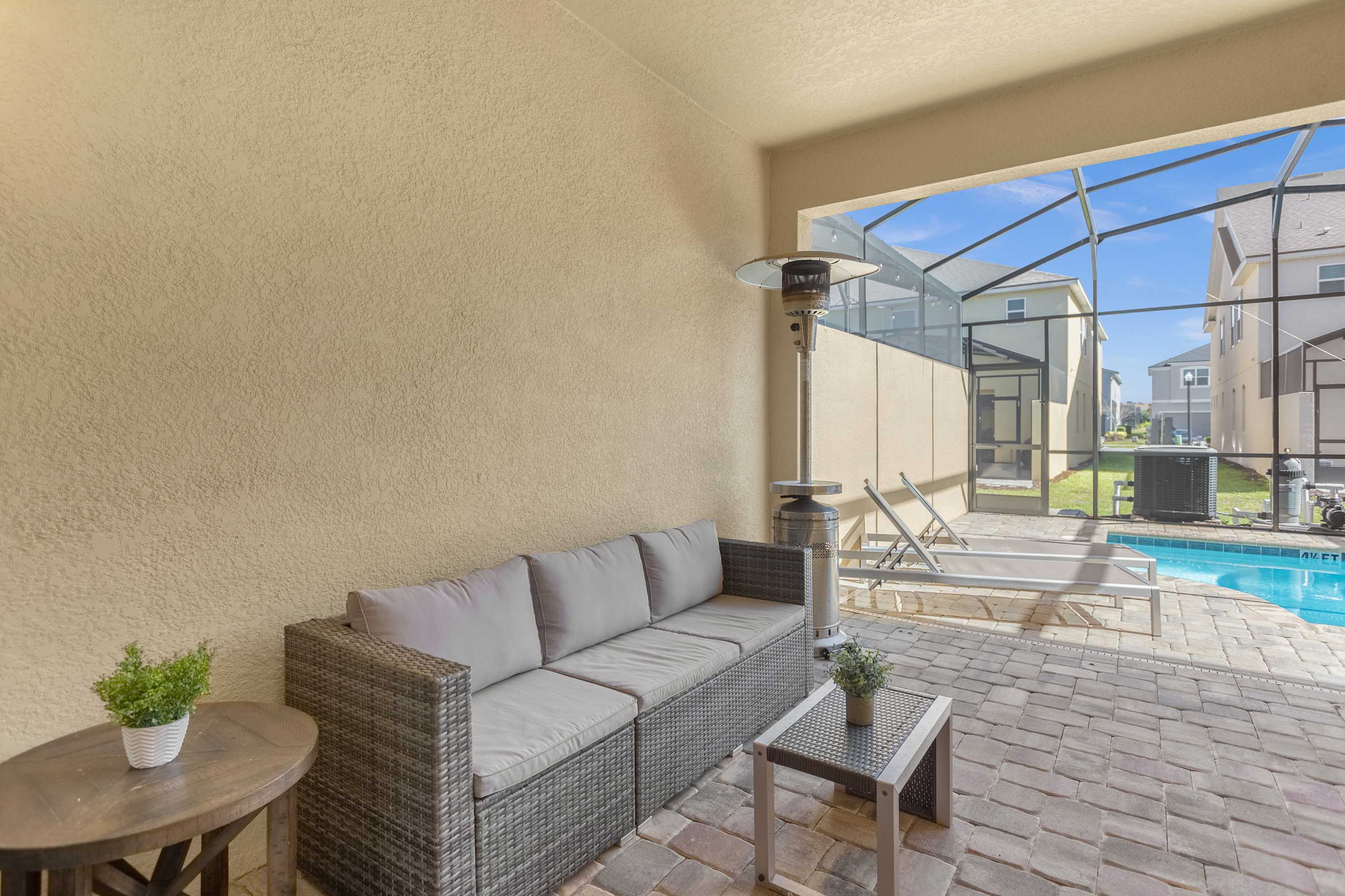 A covered patio area features a wicker sofa with gray cushions and a matching coffee table, along with a round side table. A patio heater stands nearby, overlooking a screened-in private pool area with lounge chairs.