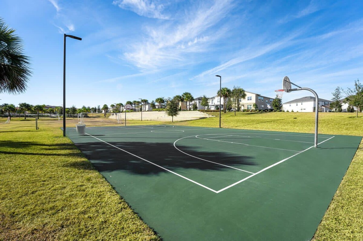 A ground-level view of a green outdoor basketball court with white boundary lines and a hoop, set on a sunny day with a clear blue sky, surrounded by green grass and distant residential homes.