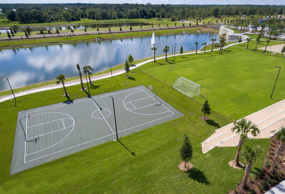 An aerial view of outdoor recreational facilities, including a green basketball court with white markings and a grassy soccer field, situated beside a serene lake with a fountain and a paved walking path.